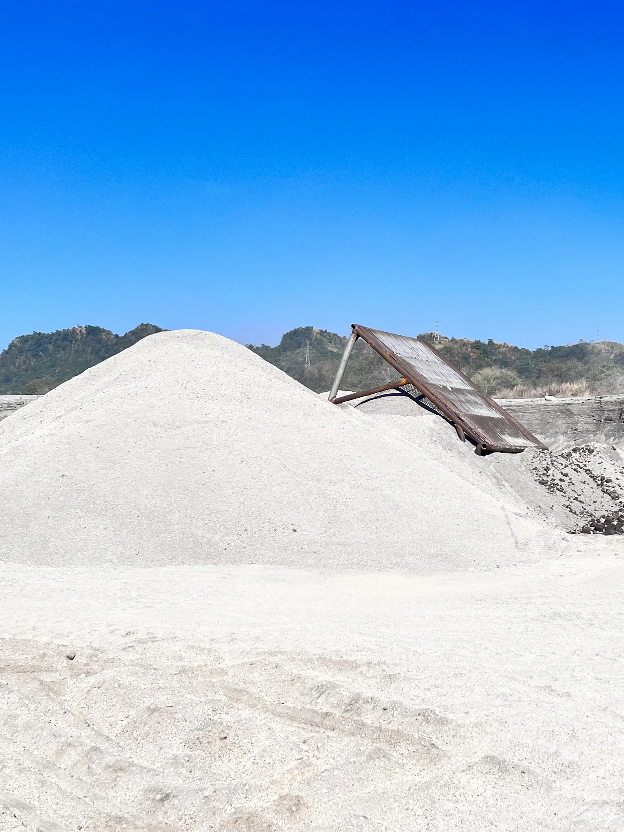 A large pile of white sand with a solar panel leaning against it, set outdoors under a clear blue sky with distant green hills in the background.