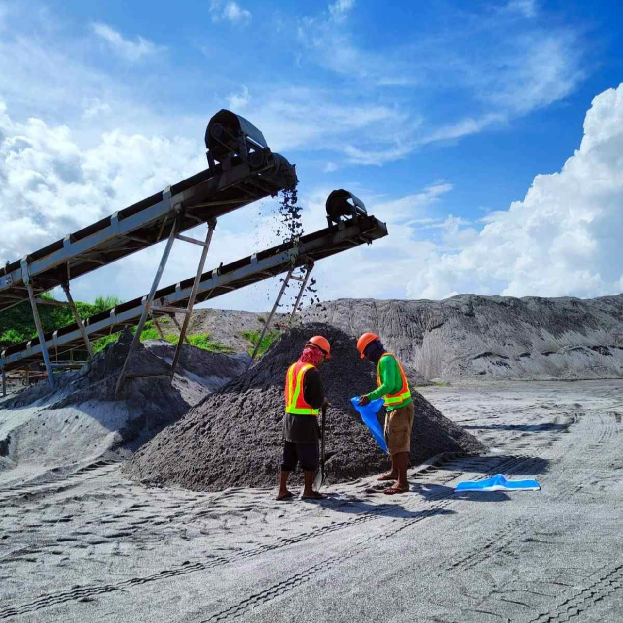 Two construction workers wearing safety helmets and vests standing next to a pile of dirt or gravel at a construction site with conveyor belts overhead and a cloudy blue sky.