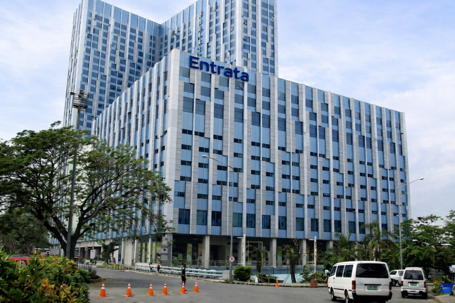 A tall modern office building with blue and white exterior panels and an entrance sign labeled 'Entrata'. Cars are parked in front and around the building, with trees and a cloudy sky in the background.