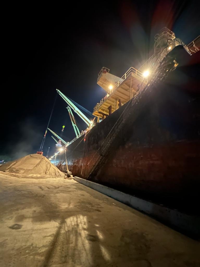 Night view of a large cargo ship docked at port, with industrial lights brightening the scene and a crane in operation, unloads or loads materials onto the ship.