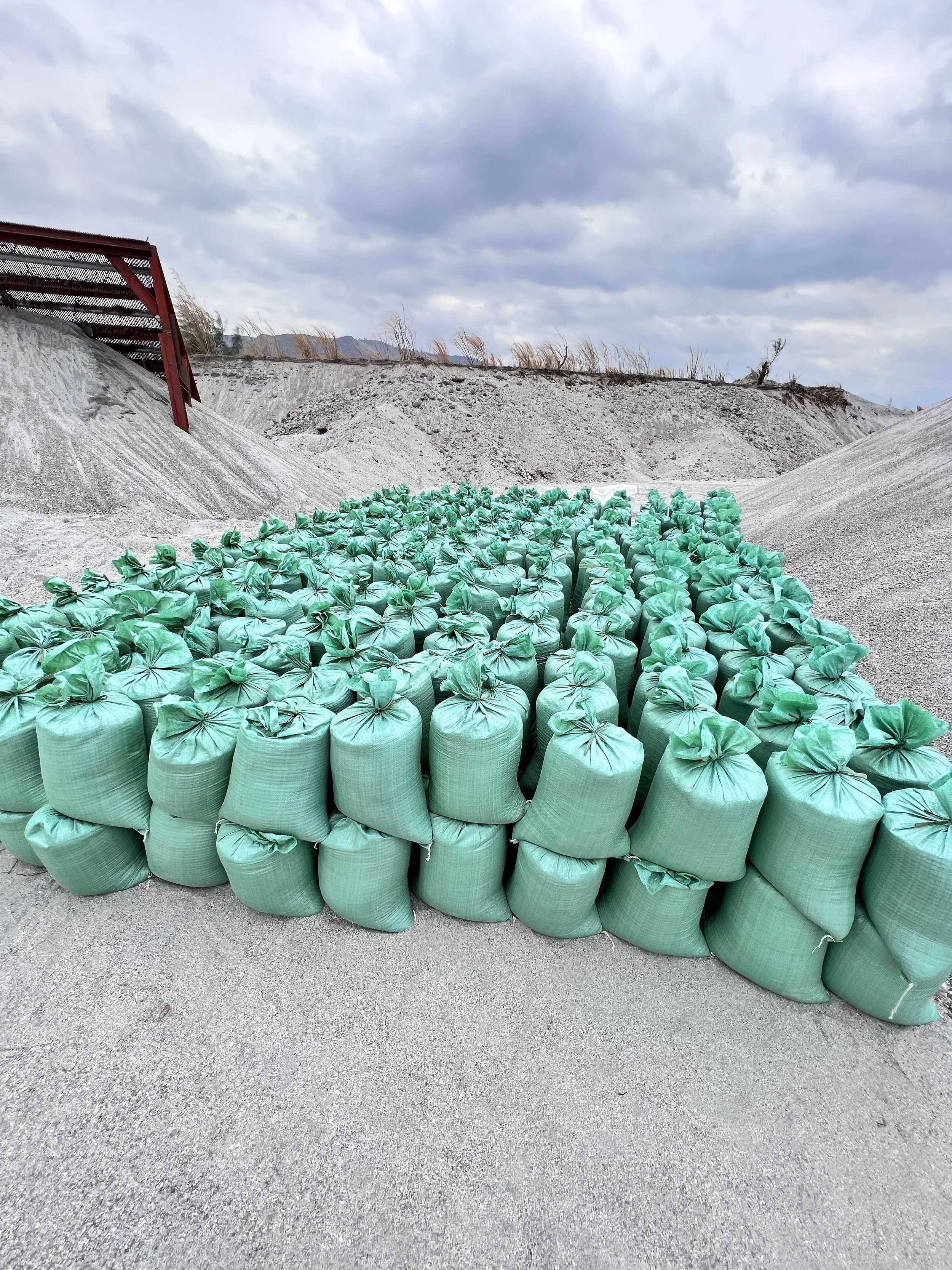 A large group of green sandbags stacked in an outdoor area with gravel, hills, and cloudy sky in the background.