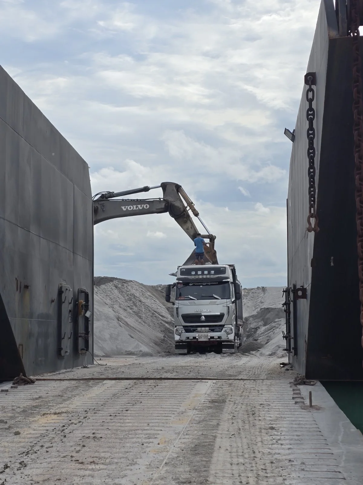 A construction scene with a large Volvo excavator lifting or moving cargo from a truck amidst a sandy or gravel landscape under a partly cloudy sky.