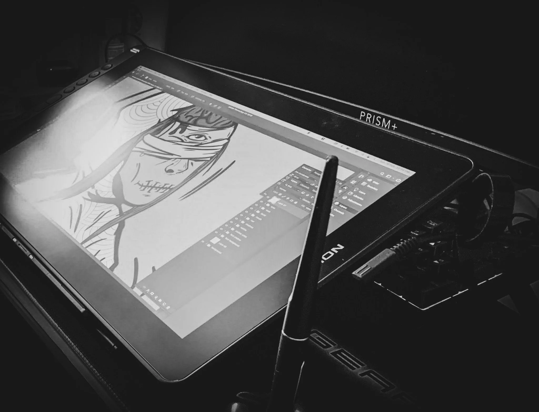 A digital drawing tablet displaying a black and white illustration of a person with an eye patch, with a stylus pen on the screen. The tablet is on a desk with some cables and a pen holder nearby. The screen shows drawing software, and the device has a label "PRISM+".