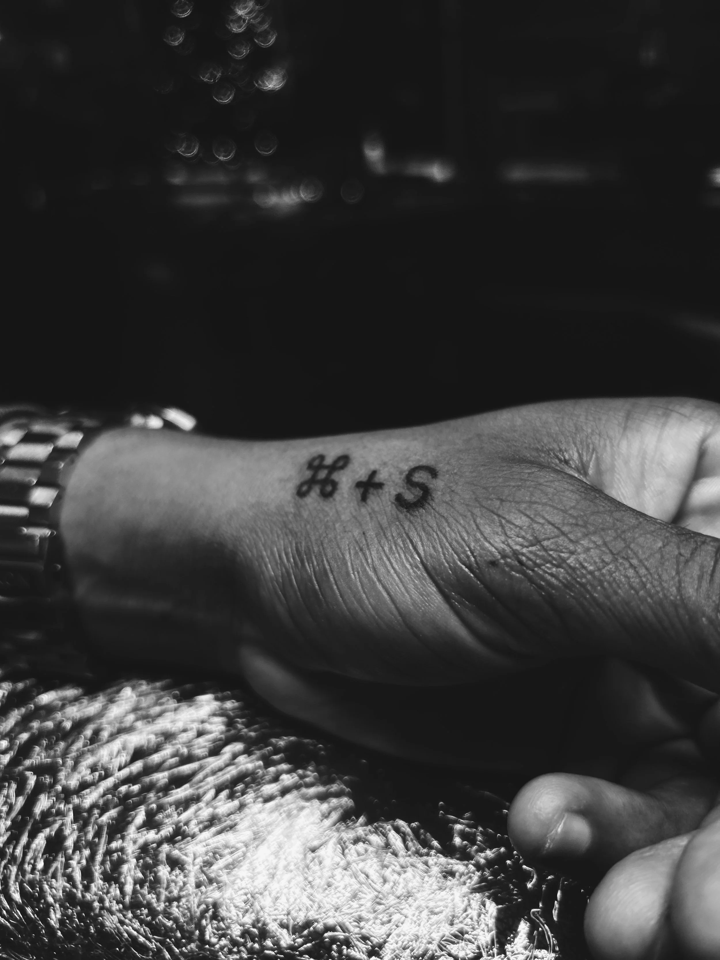 Black and white photo of a hand with a tattoo of the keyboard command symbols '⌘ + S' on the wrist, wearing a watch.