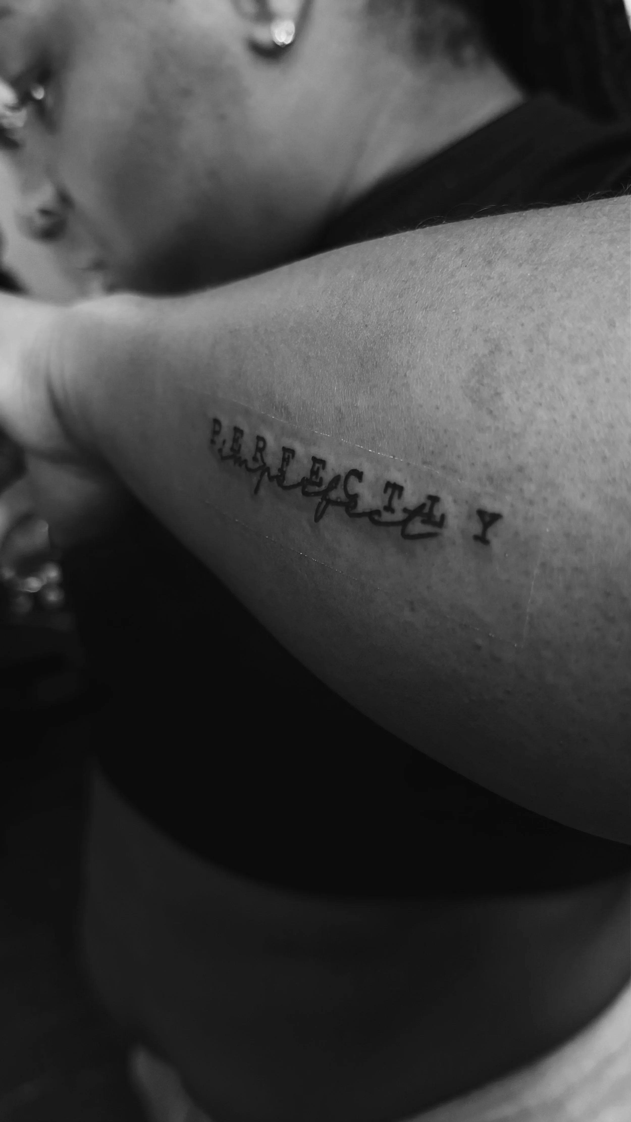 Close-up of a person's arm with a tattoo of the word "BELIEVE" in black ink, some of which is in a cursive font, and the person's face partially visible in the background.