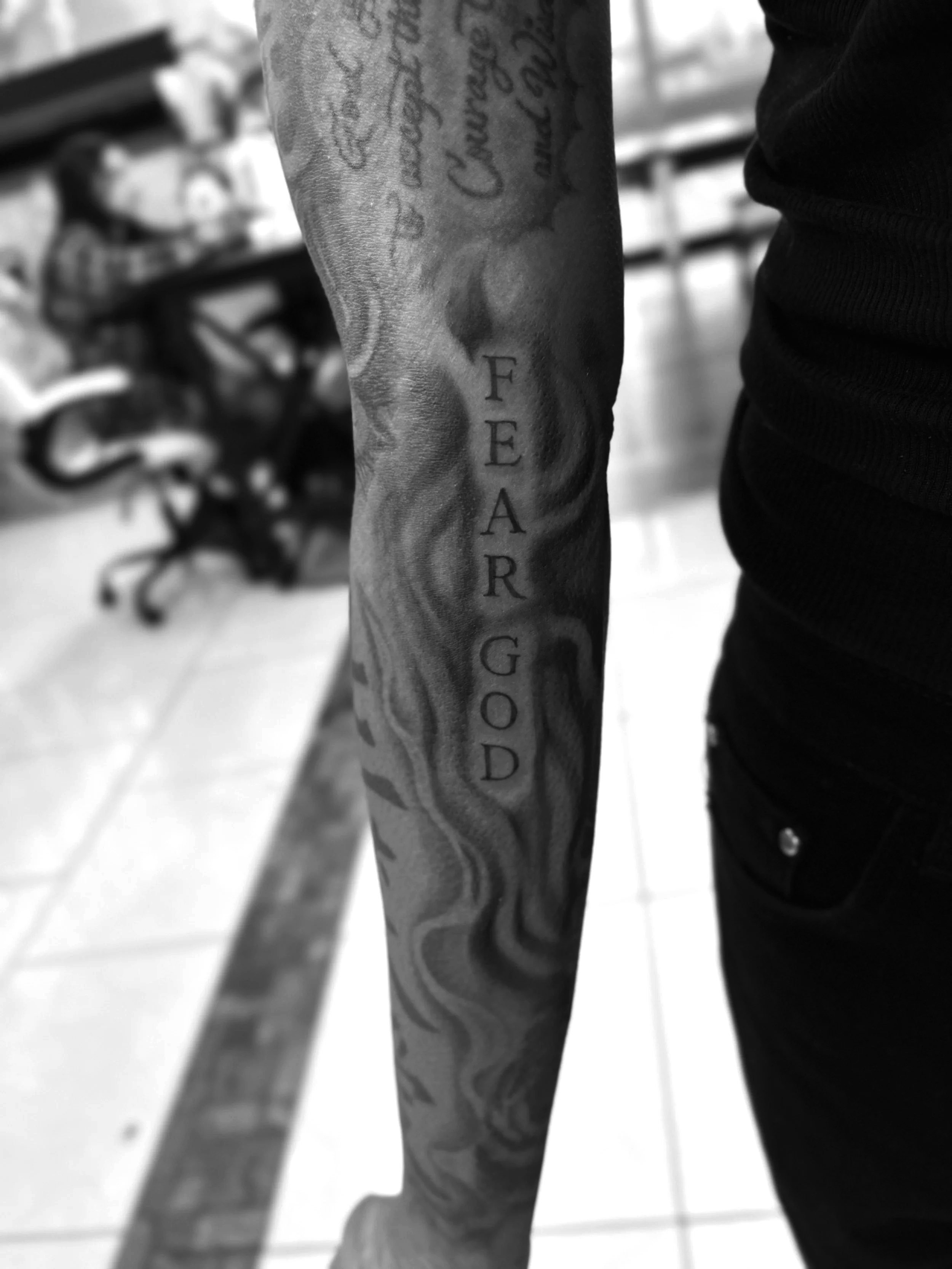 Tattooed forearm with the phrase 'FEAR GOD' written vertically in capital letters on a background of flowing smoke or flames.