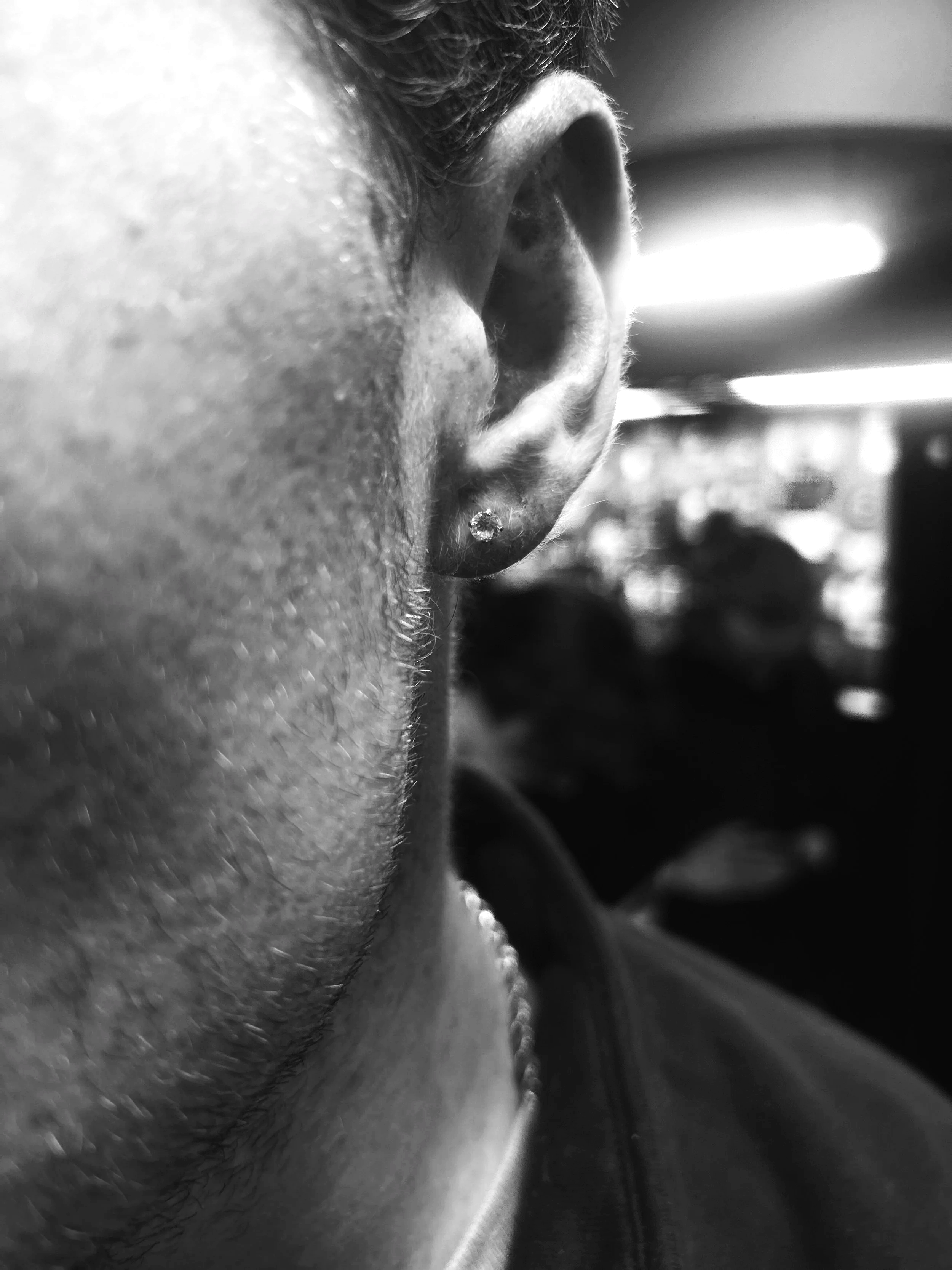 Close-up black and white photograph of a person's ear with a small diamond earring, with a blurred background of indoor lighting and people in a room.