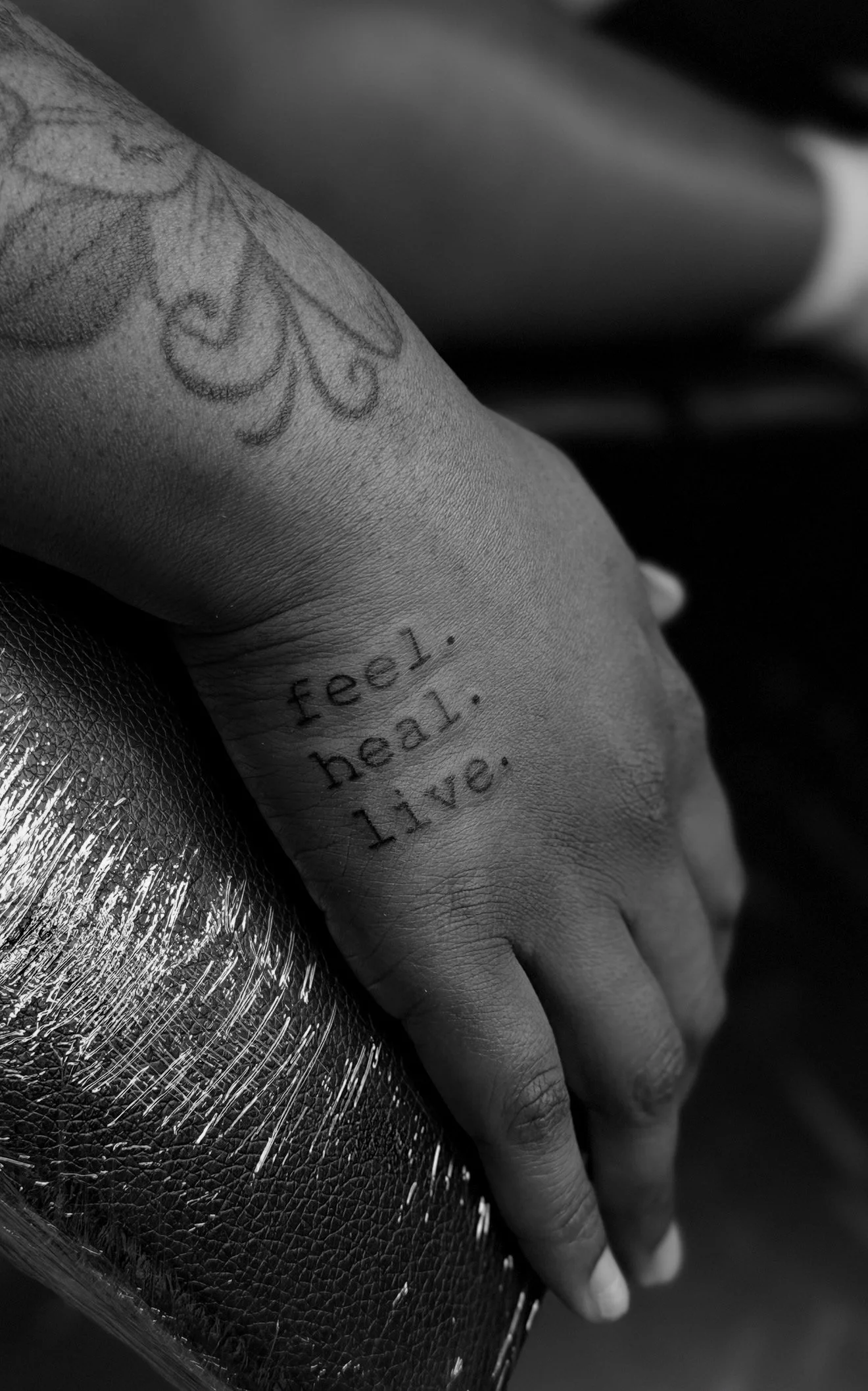 A close-up of a person's hand with a tattoo that says "feel, heal, live." on the wrist, resting on textured fabric in black and white.