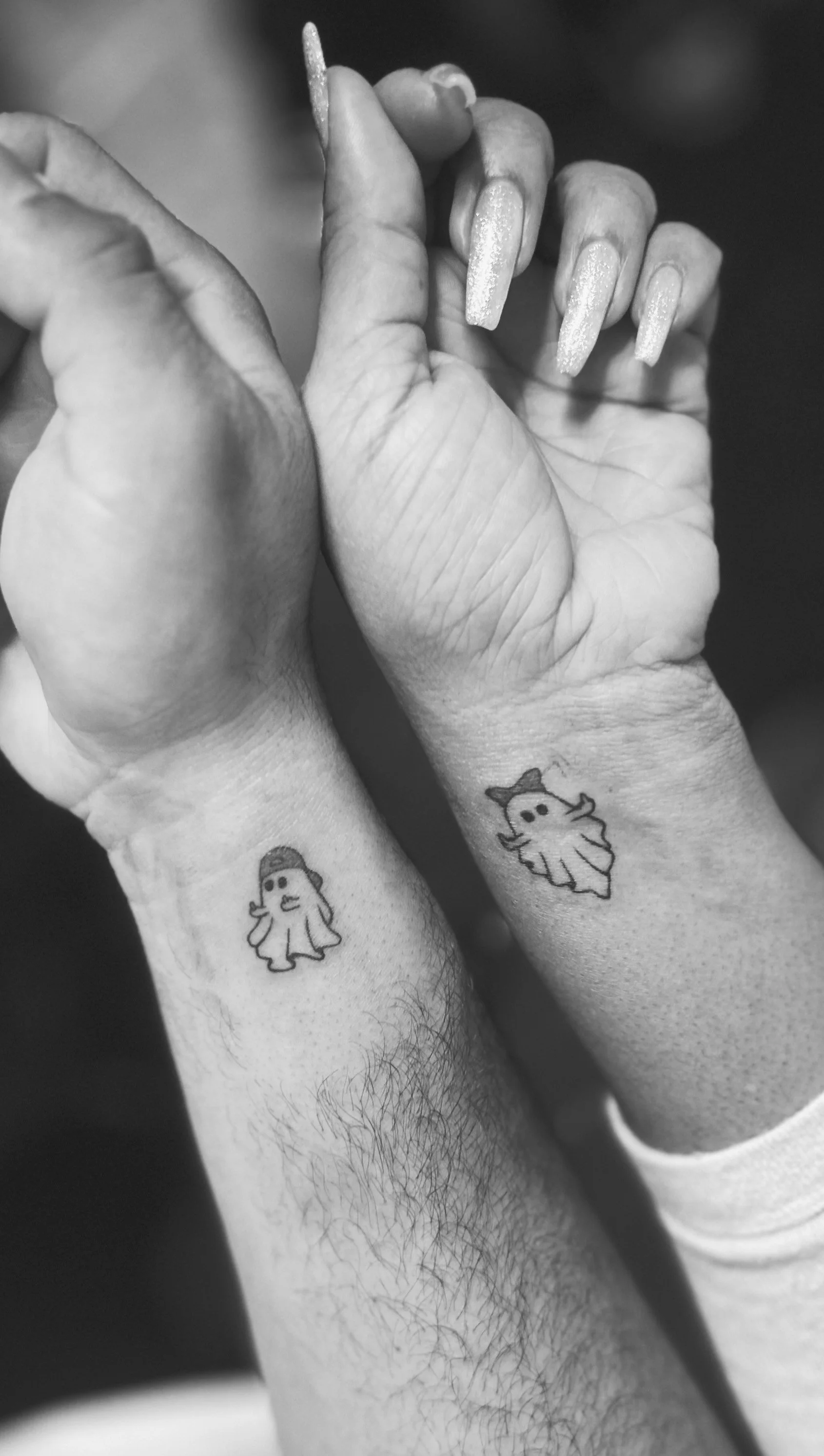 Two hands with matching tattoos of cartoon ghosts on their wrists, one wearing a baseball cap, in black and white.