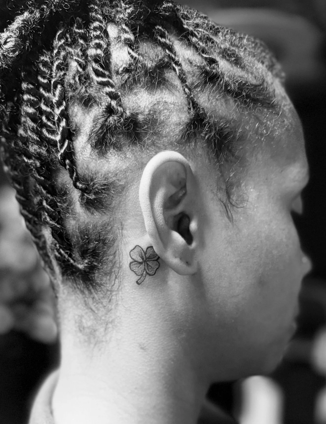 Close-up of a person's head with braided hair, a tattoo of a four-leaf clover behind the ear, and short hair on the neck in black and white.