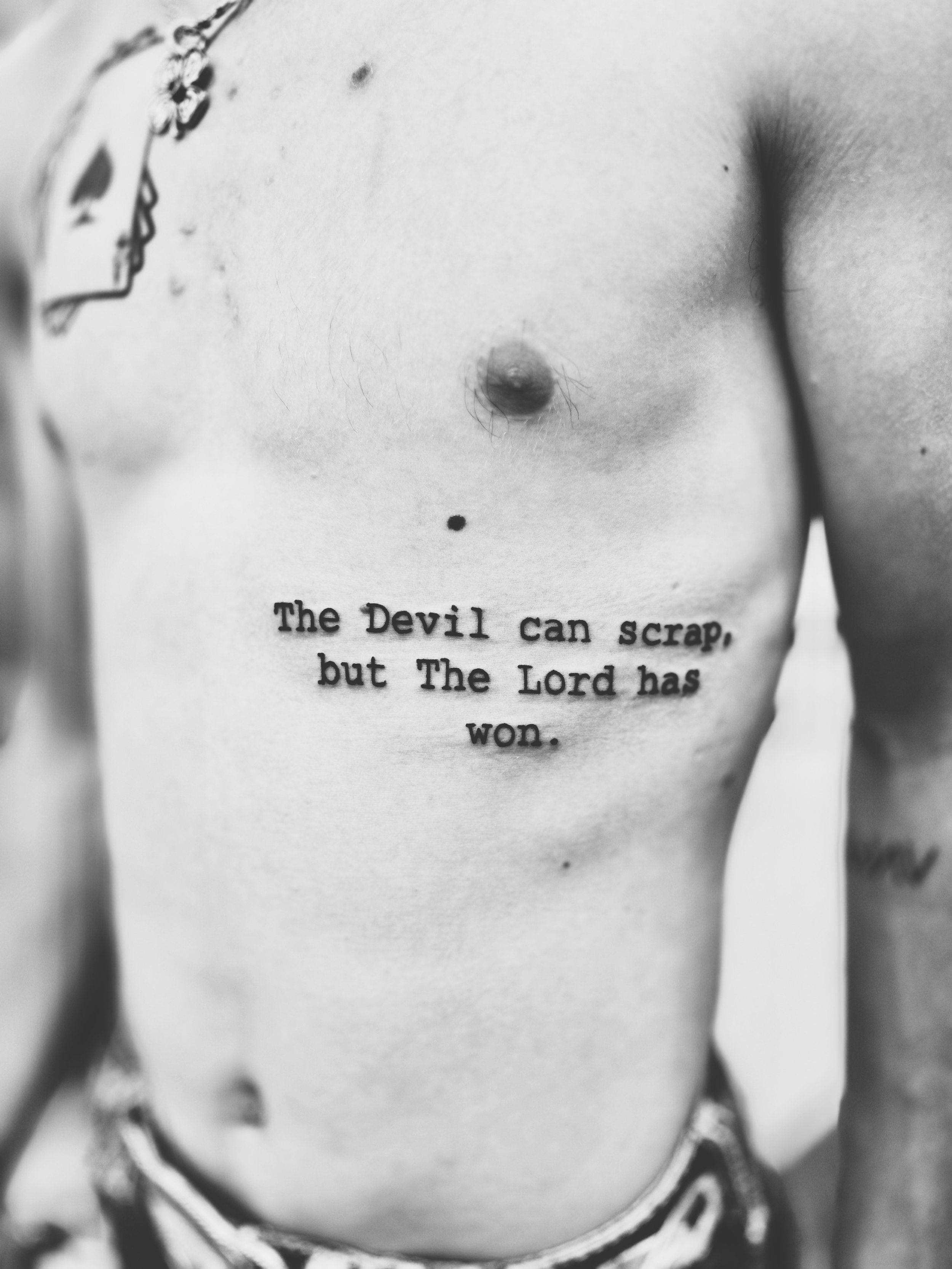 Black and white tattoo on a person's torso reading 'The Devil can scrap, but The Lord has won.'