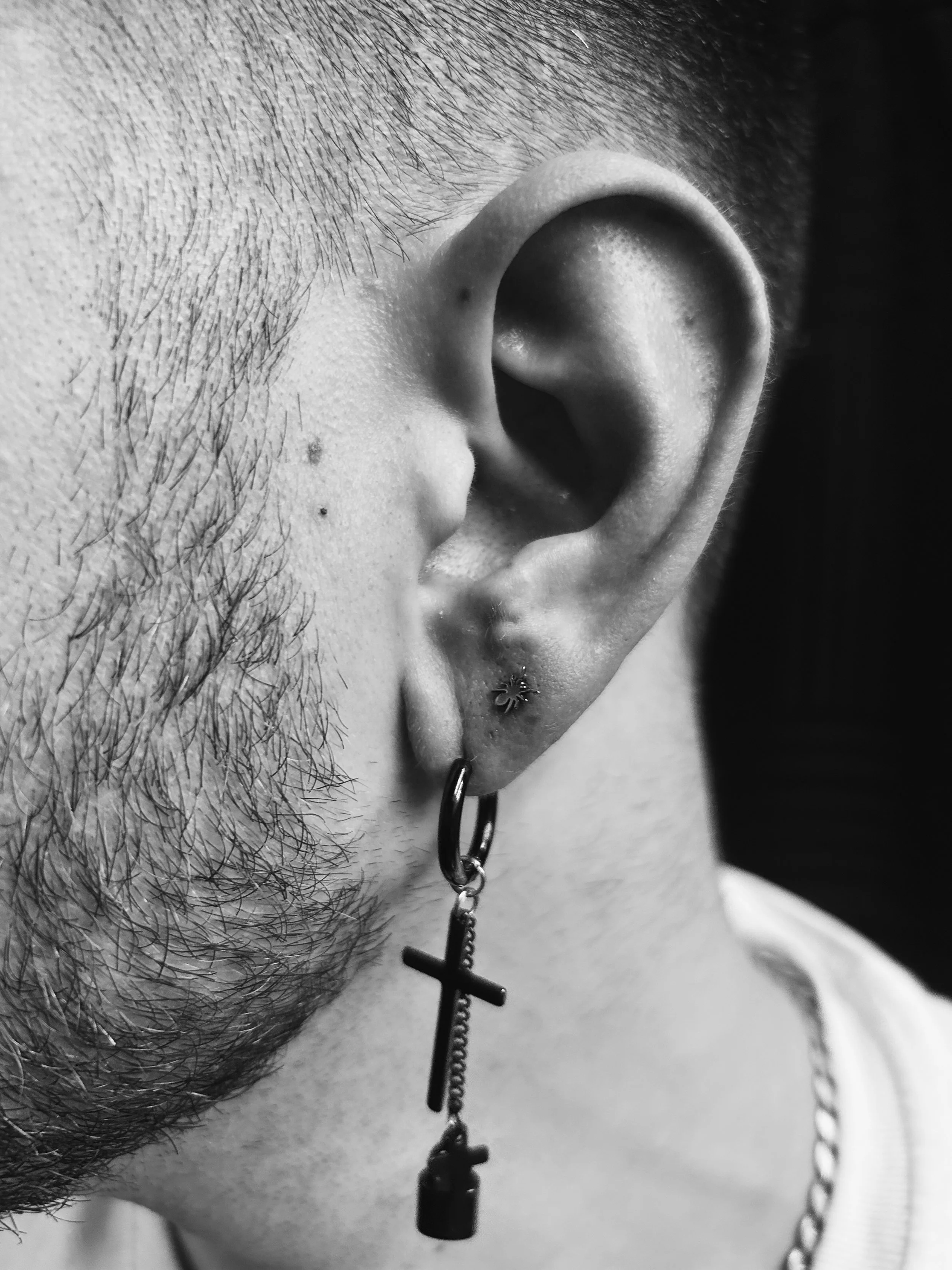 Close-up of a person's ear with multiple piercings, including a small hoop with a keychain charm and a small star-shaped earring, and some facial hair visible.