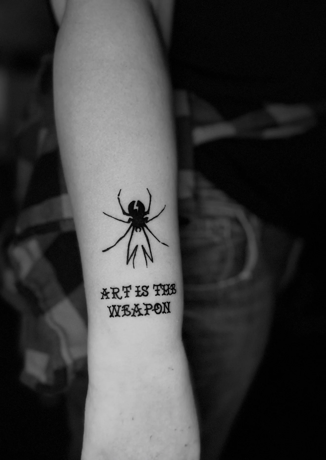 Tattoo of a spider with a message below that reads 'Art is the Weapon' on a person's forearm.