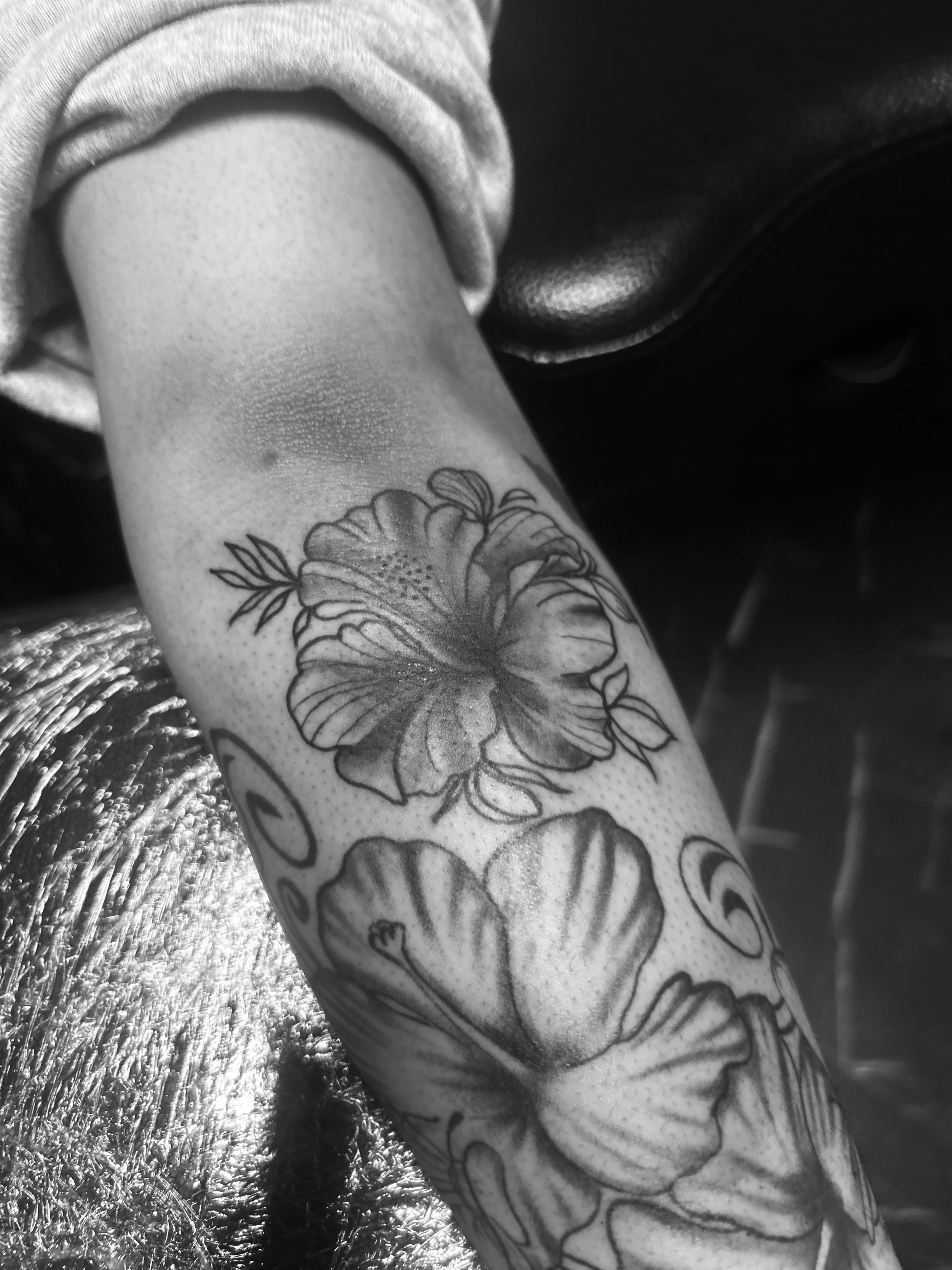 Black and white photo of a person's lower arm with a floral tattoo featuring hibiscus flowers, leaves, and other botanical elements.