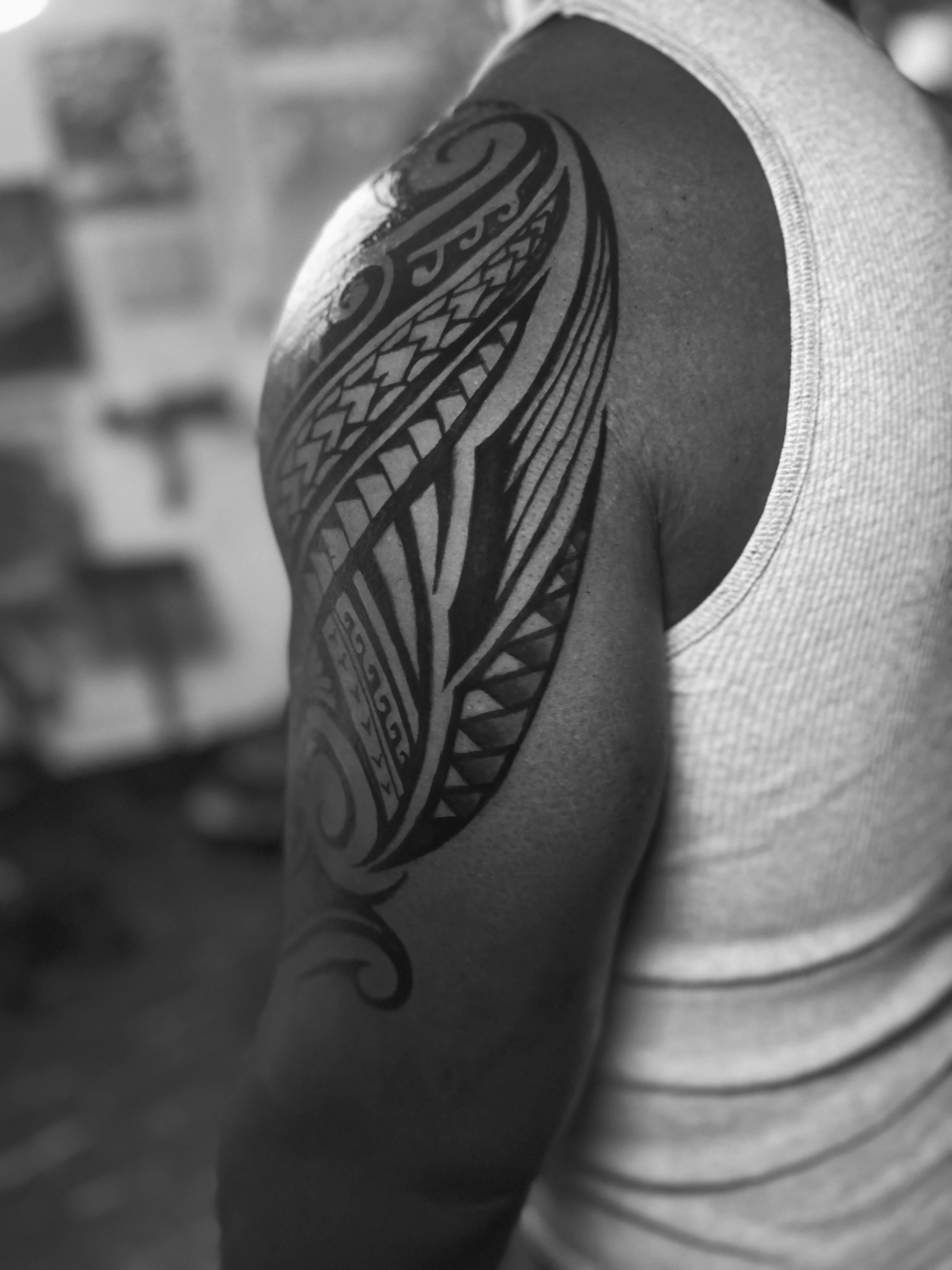 Close-up of a person's shoulder with a Polynesian-style tribal tattoo, wearing a sleeveless shirt.