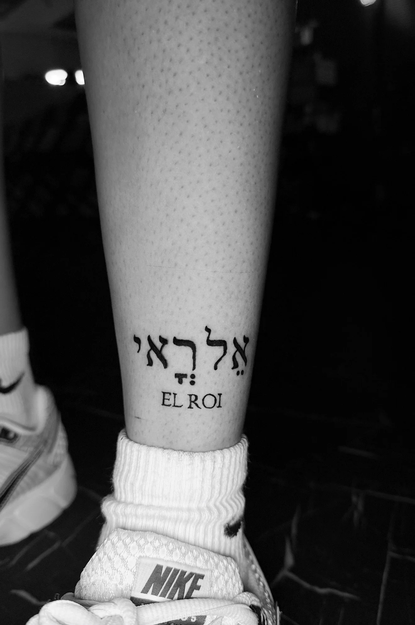 Close-up of a person's leg with a tattoo in Hebrew and English that reads 'EL ROI.' The leg is wearing a white sock and a Nike sneaker.