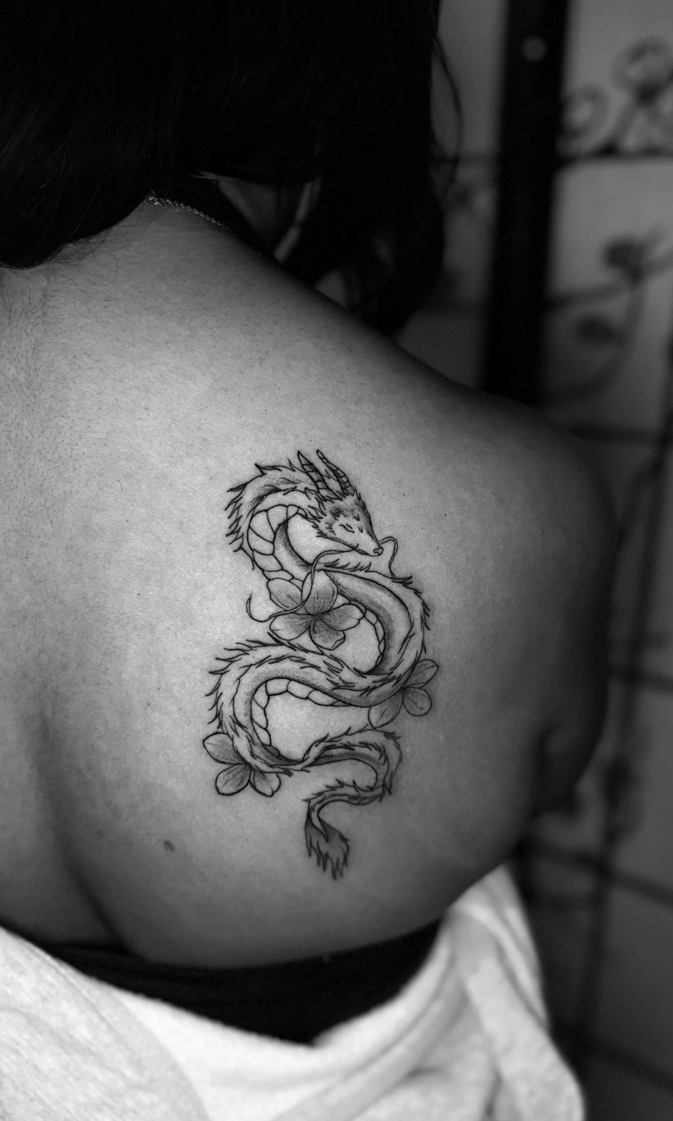 Black and white tattoo of a dragon intertwined with flowers on a person's shoulder.