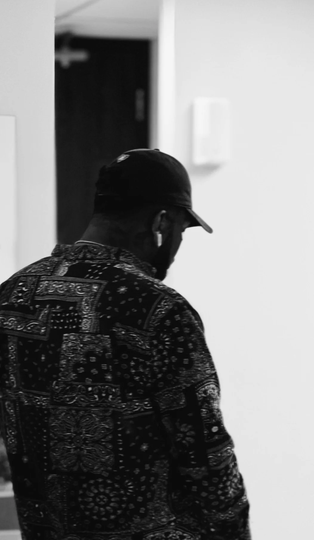 A man with a cap and patterned jacket standing with his back turned, in black and white.