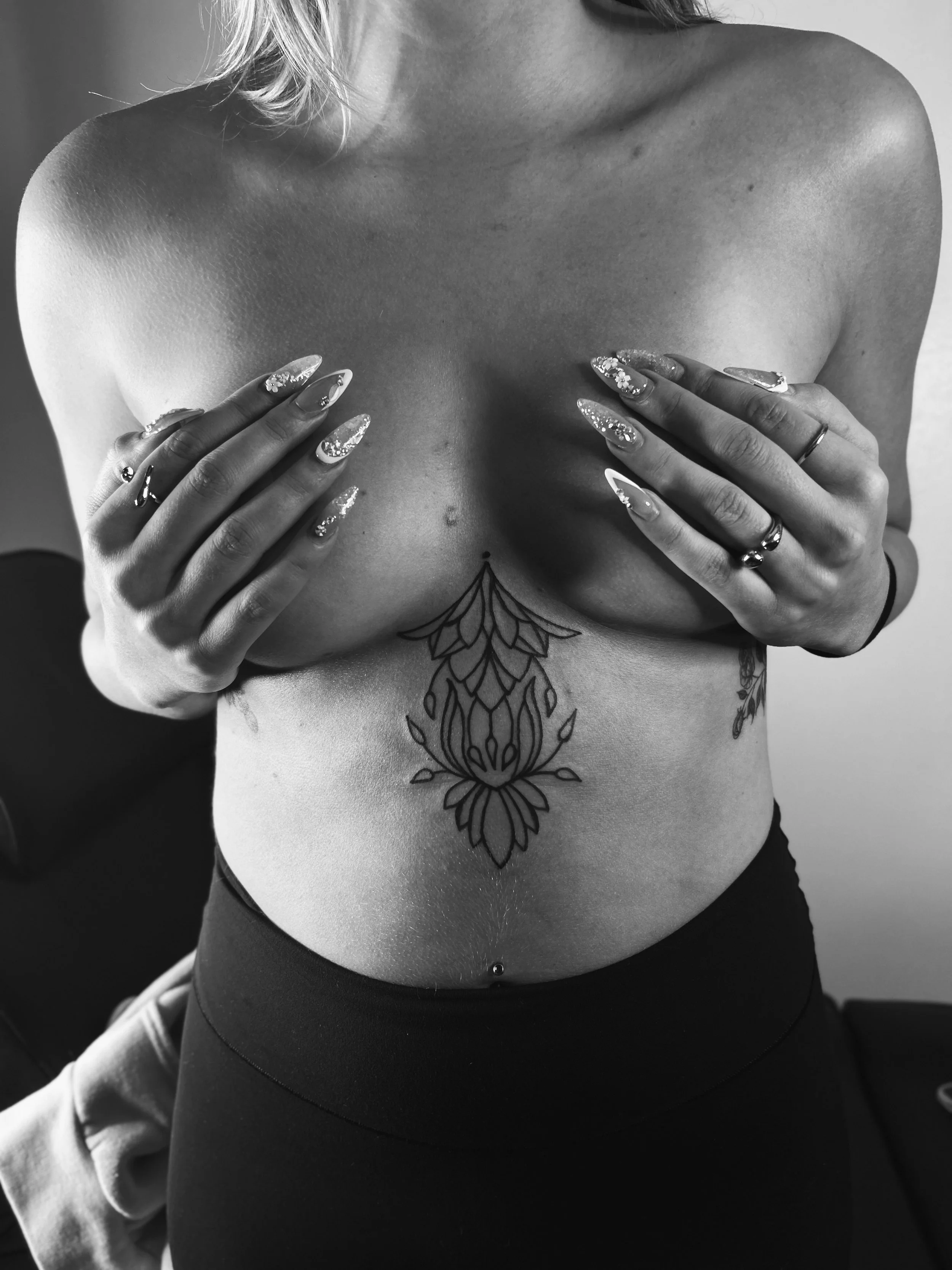 A woman with light-colored hair covering her breasts with her hands. She has long, decorated nails and multiple rings on her fingers. She has a large floral tattoo on her abdomen and a belly button piercing. The photo is in black and white.