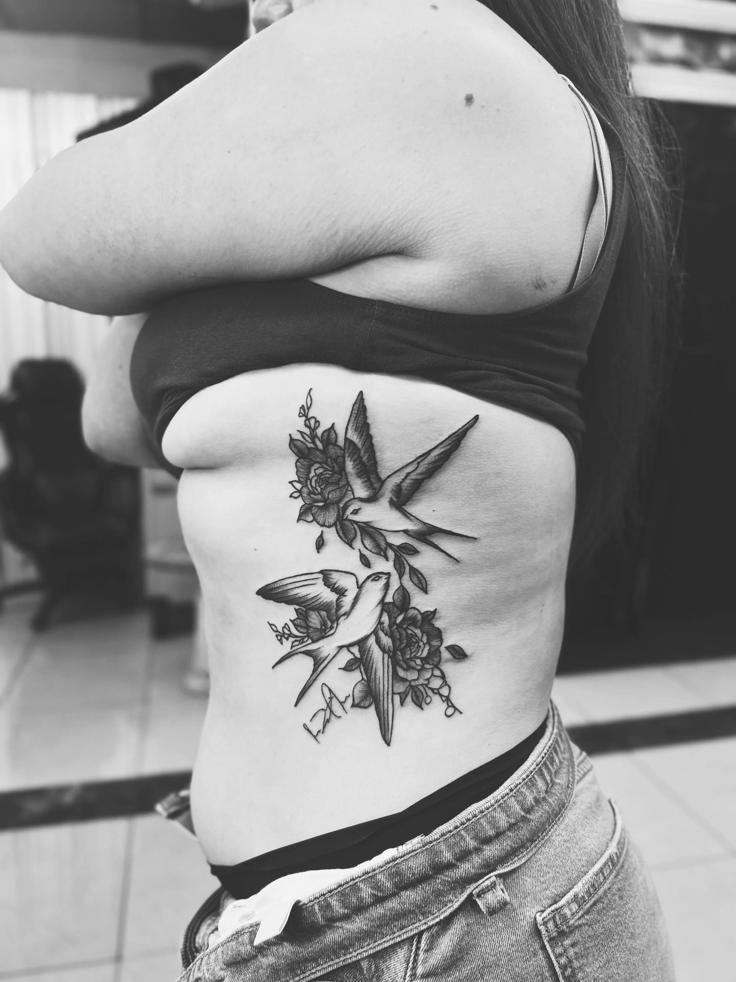 Black and white photo of a woman's side torso showing a tattoo of two hummingbirds and flowers.