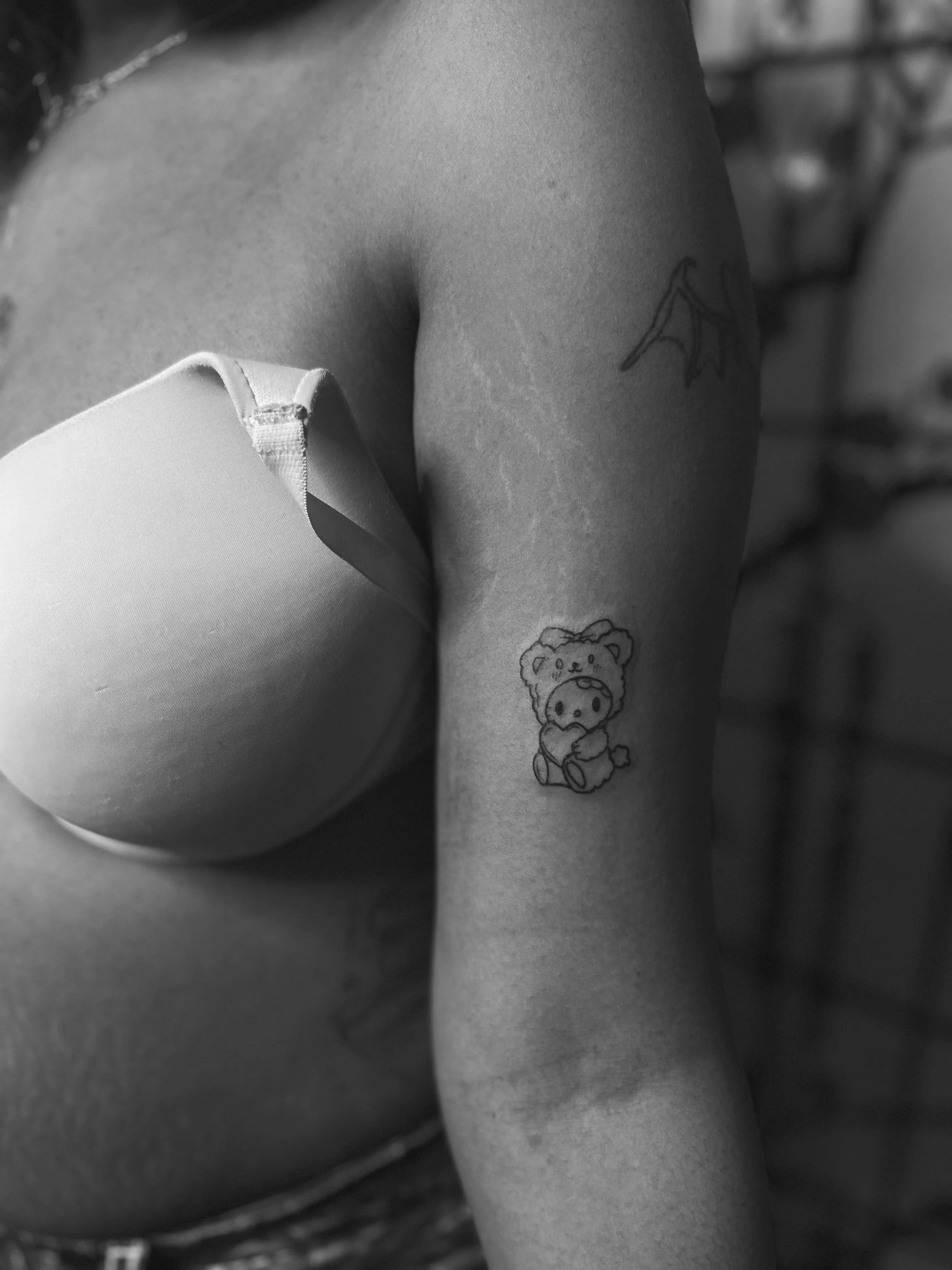 Close-up of a person's upper body, showing two tattoos on the arm: one of a bear's face and another of a cute bear holding a star. The person is wearing a strapless top.