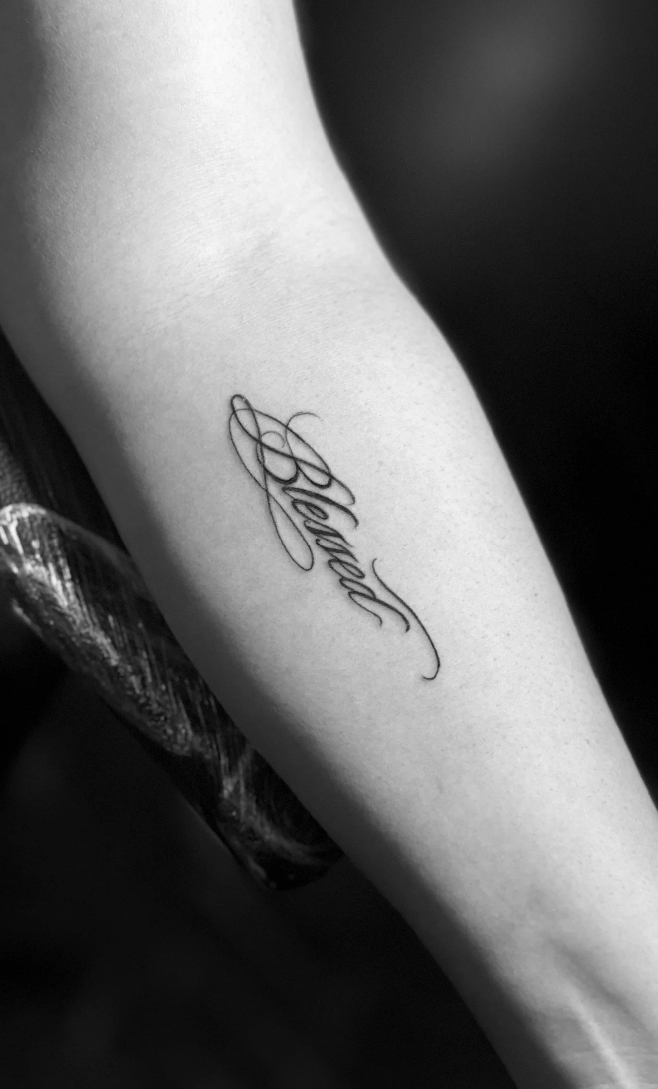 Black and white photo of a person's arm with a tattoo that says 'Blessed' in cursive script.