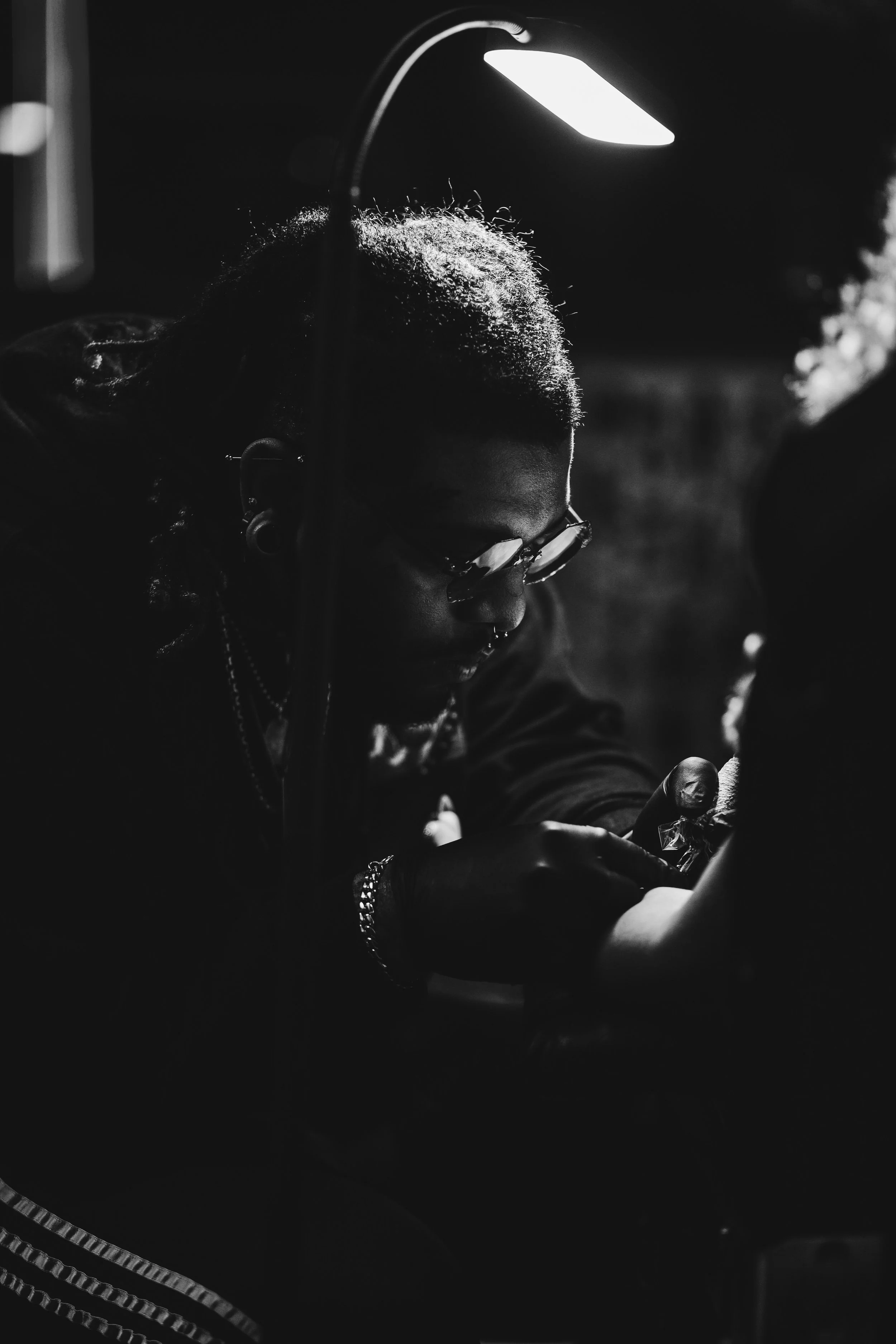 A person with glasses and jewelry getting a tattoo under a bright lamp in a tattoo studio, black and white photo.