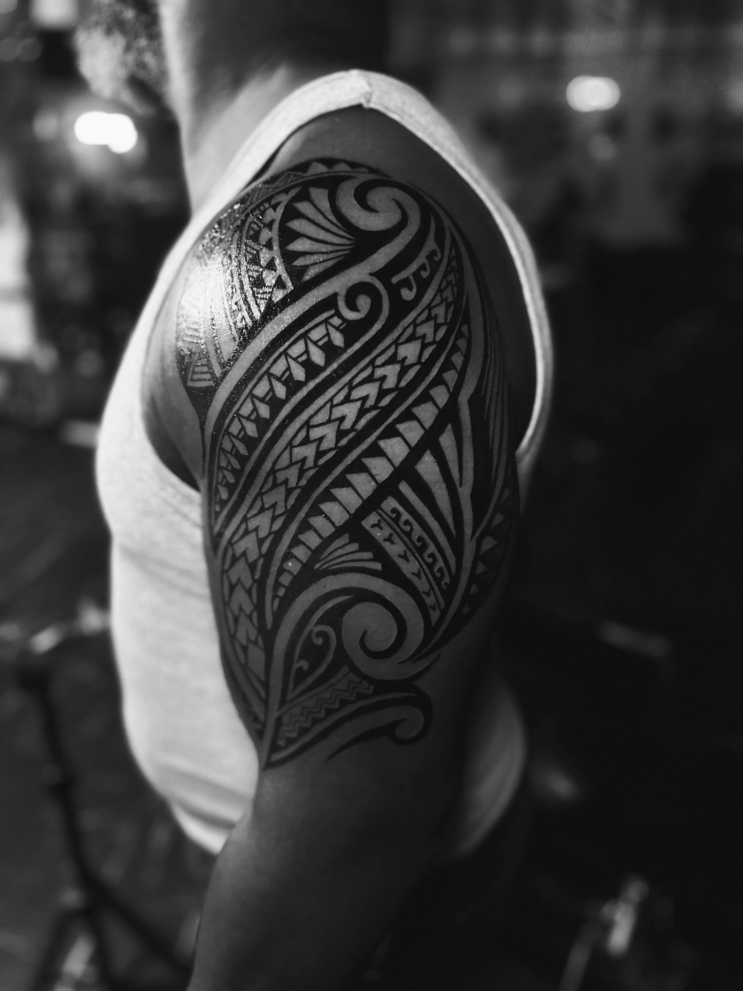 A person with a Polynesian-style tattoo on their shoulder and upper arm, wearing a sleeveless shirt, photographed in black and white.
