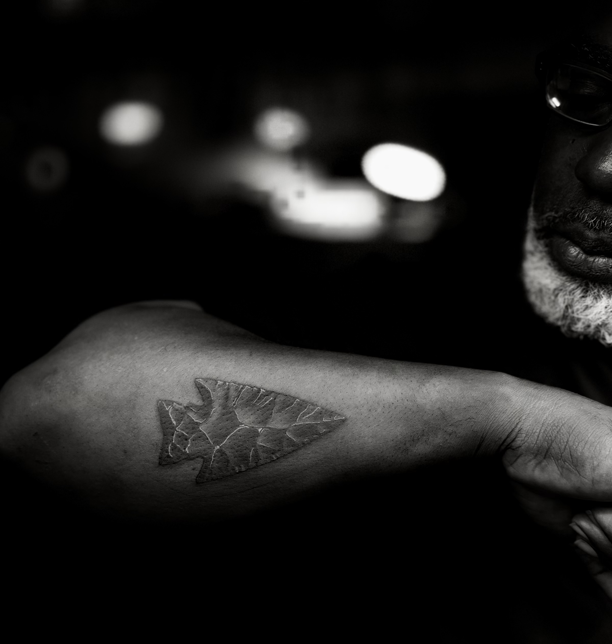 Close-up of a person's arm with a tattoo of a leaf, with a person partially visible in the background in a black and white photo.