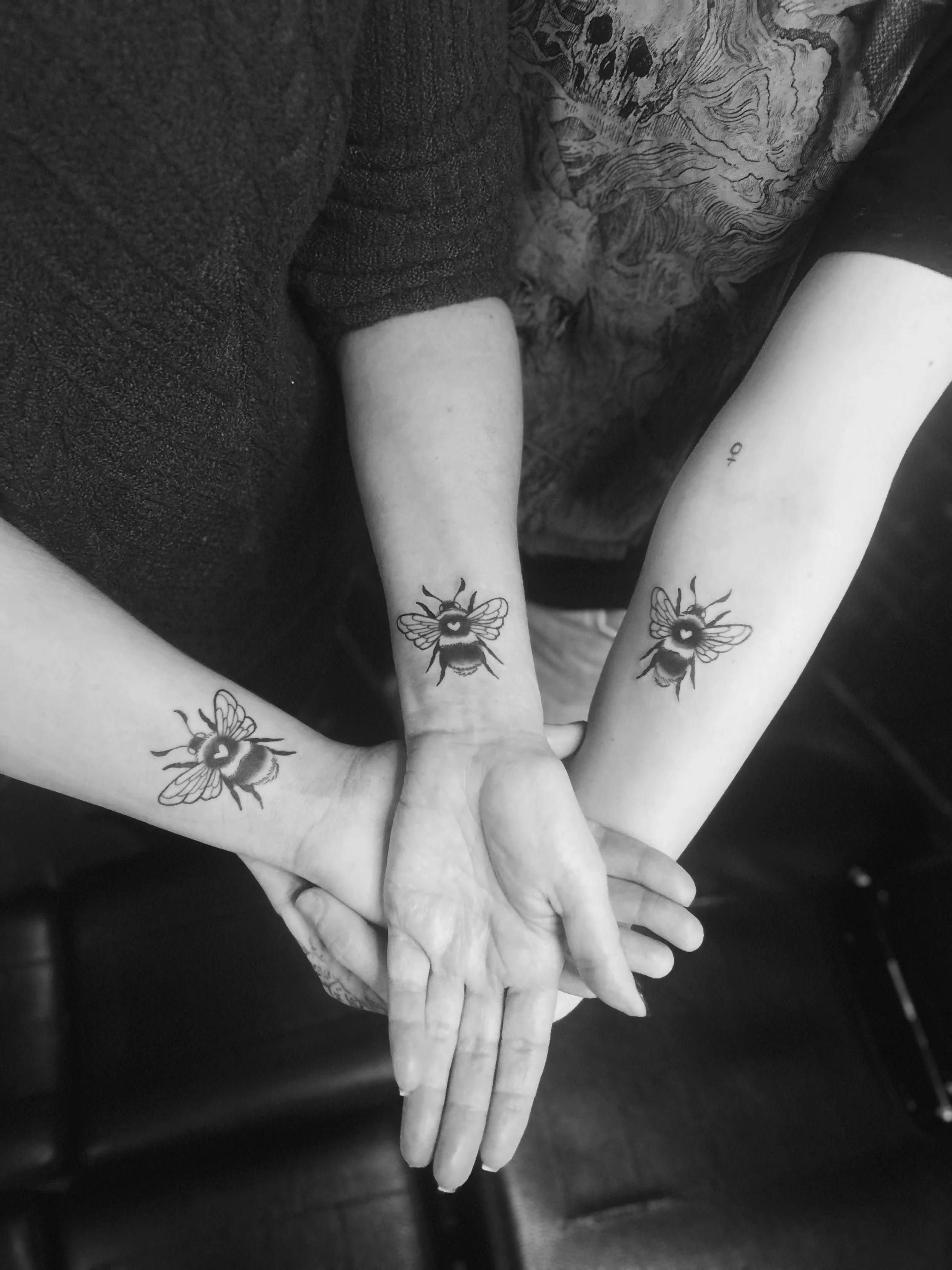 Three hands with matching bee tattoos overlapping each other, against dark and textured background.