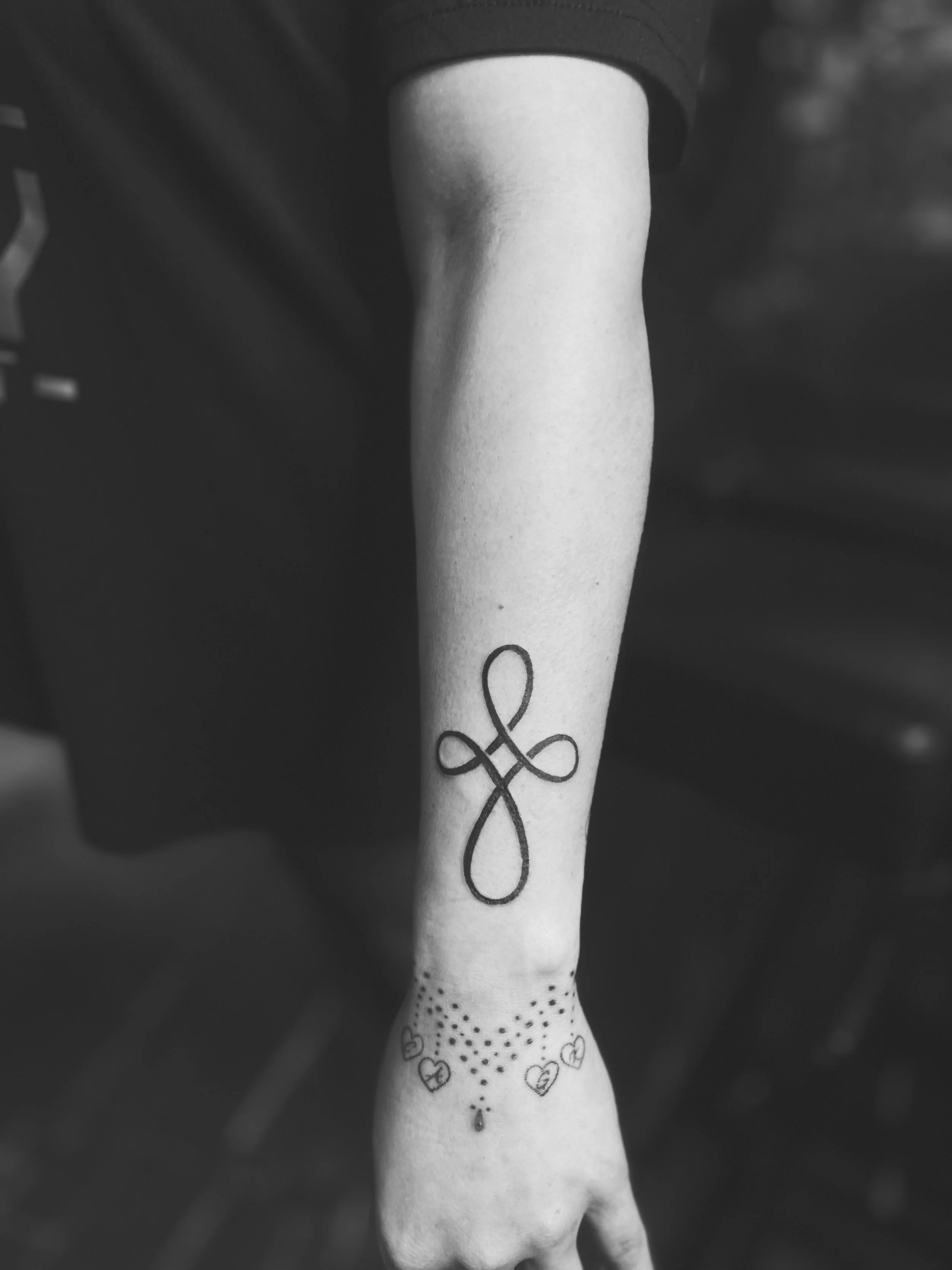 Black and white photo of a person's forearm tattoo, featuring a stylized cross with loops and small hearts and dots around it.