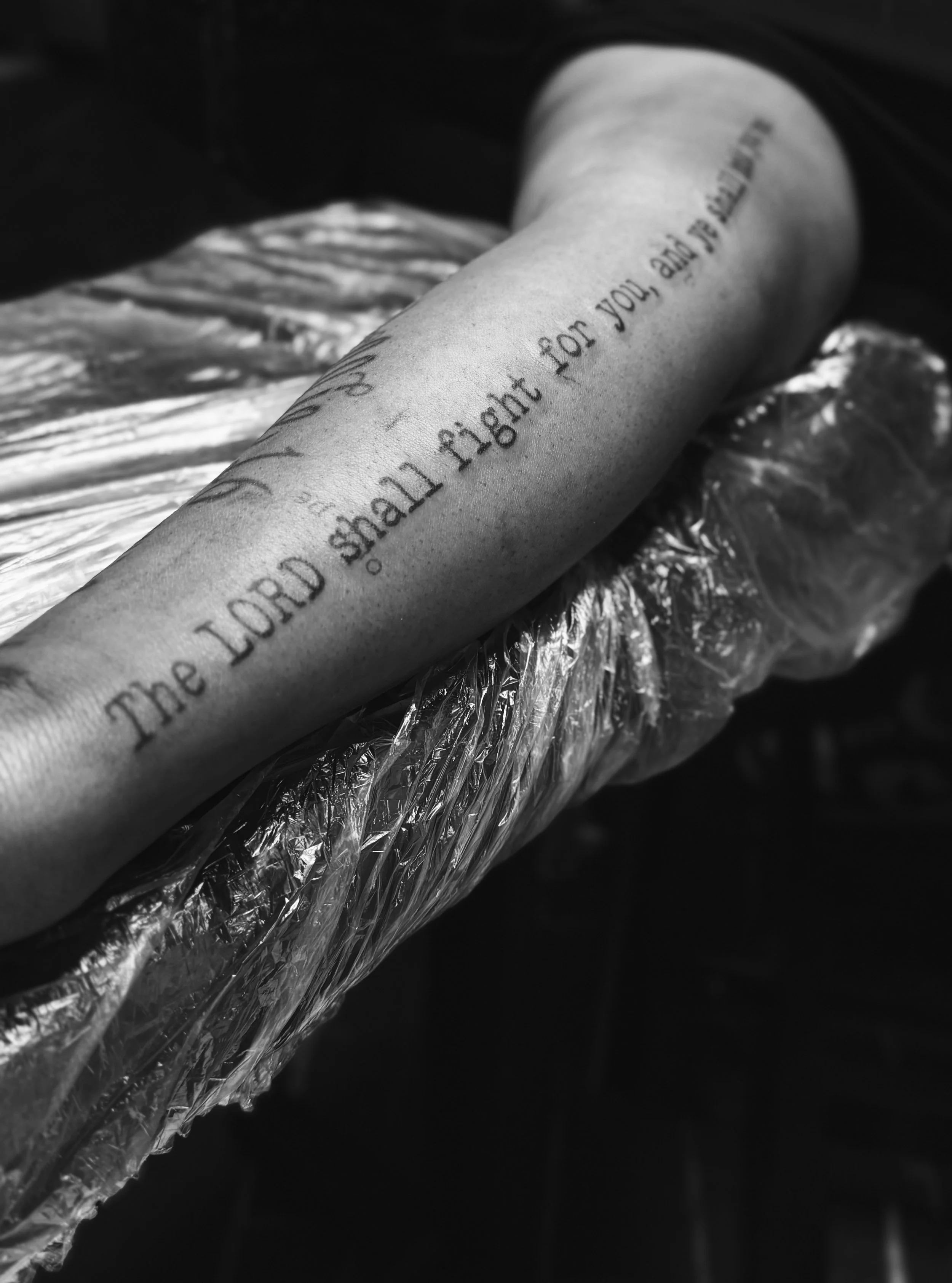 A tattoo on a person's arm with the words 'The Lord shall fight for you, and ye shall hold your peace, and be still.' The arm is resting on a surface covered with clear plastic.