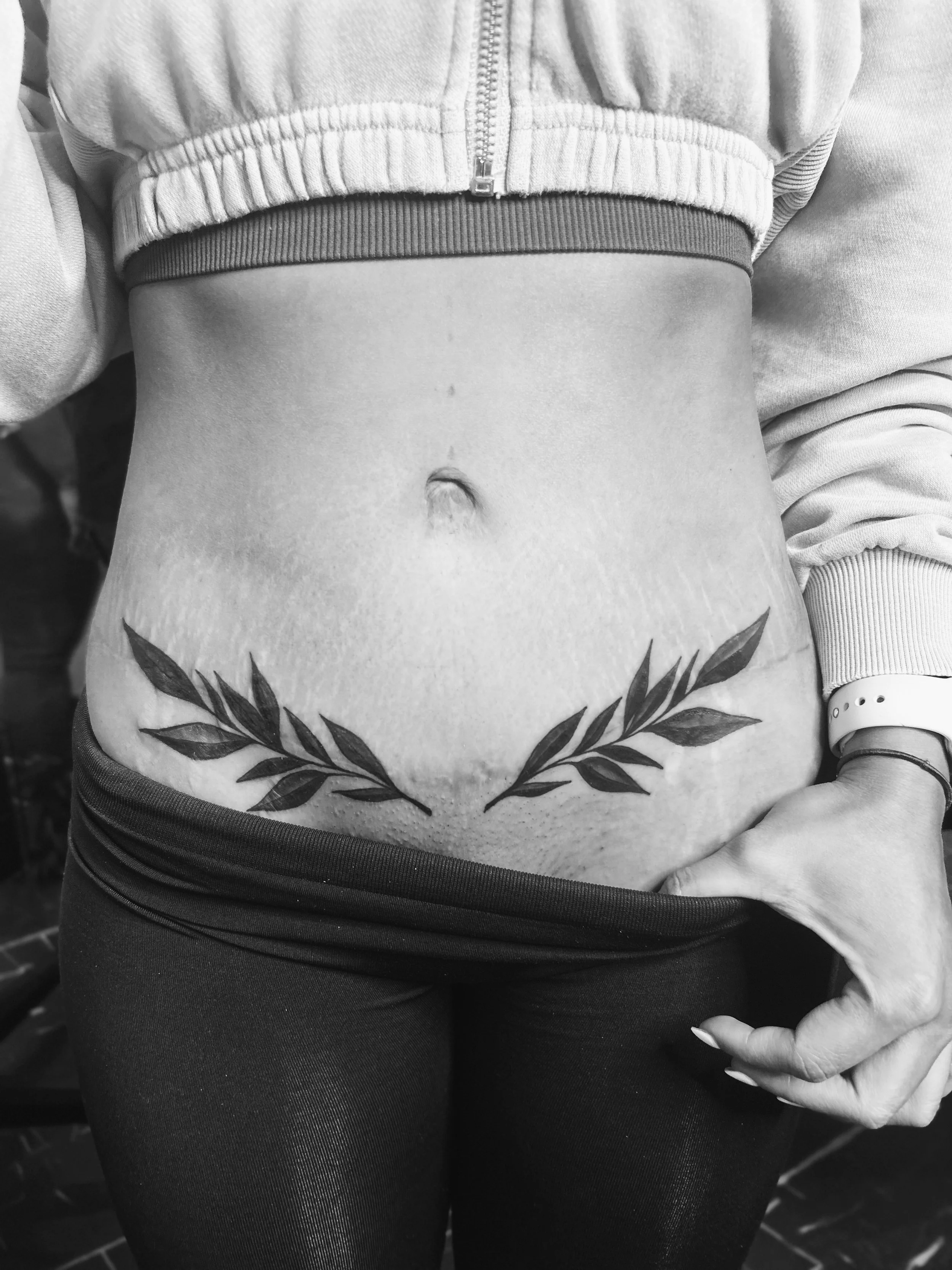Black and white photo of a person's lower torso, featuring a tattoo of laurel branches below the belly button, with one hand resting by their side and a smartwatch on their wrist.