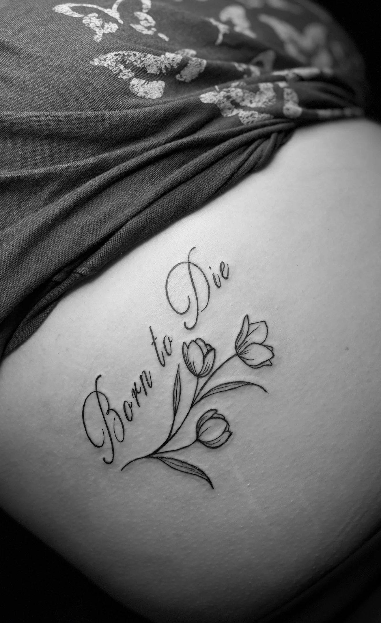 Tattoo on skin that reads 'Born to Die' with a drawing of three flowers beneath the text, partly covered by dark fabric.