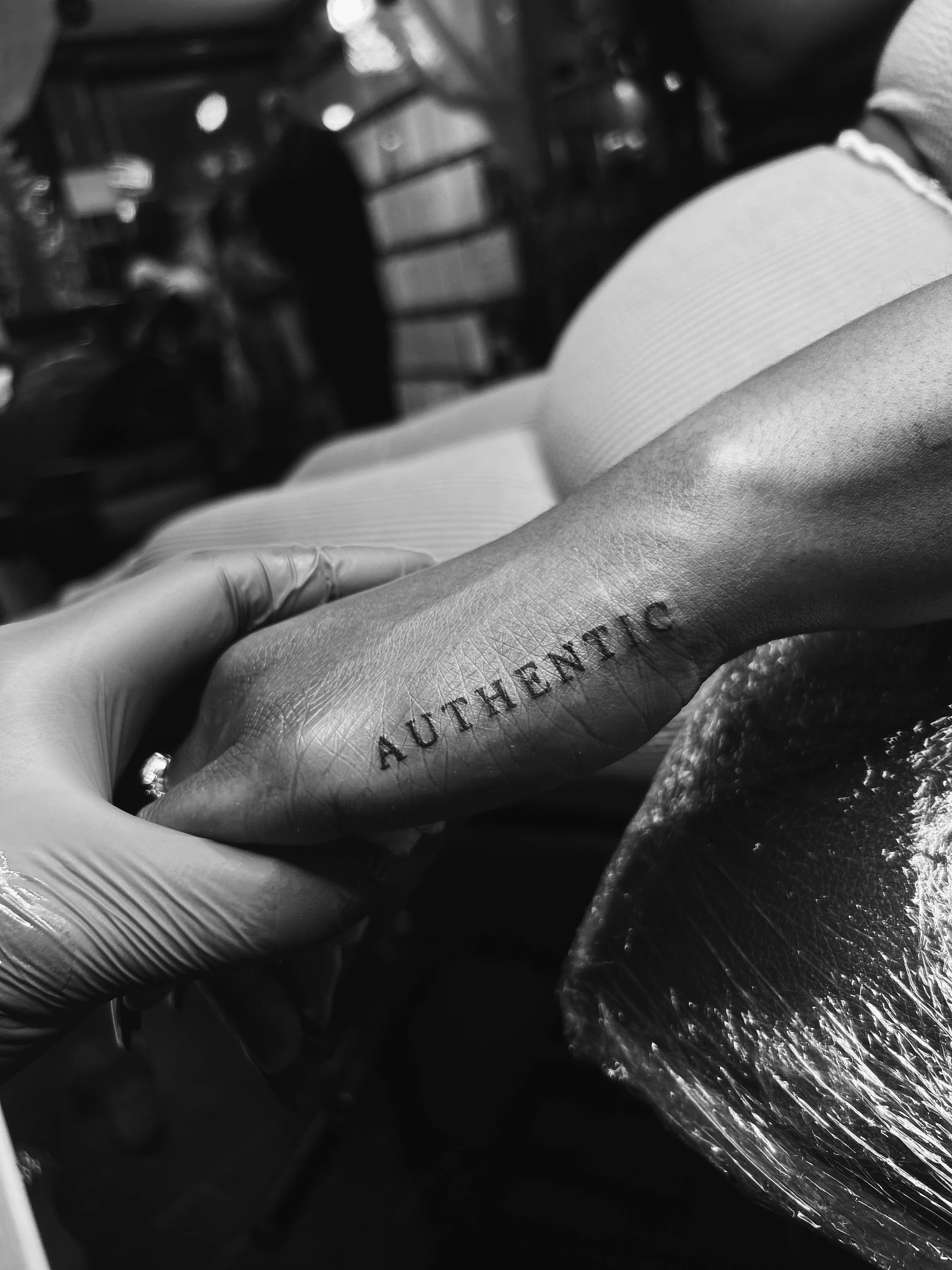 Close-up of a tattoo that reads 'AUTHENTIC' on a person's finger or hand, with a person in the background at a tattoo studio.