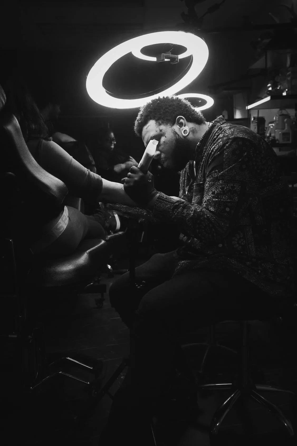 A person with large gauge earrings getting tattooed by an artist in a dimly lit studio with neon ceiling lights.
