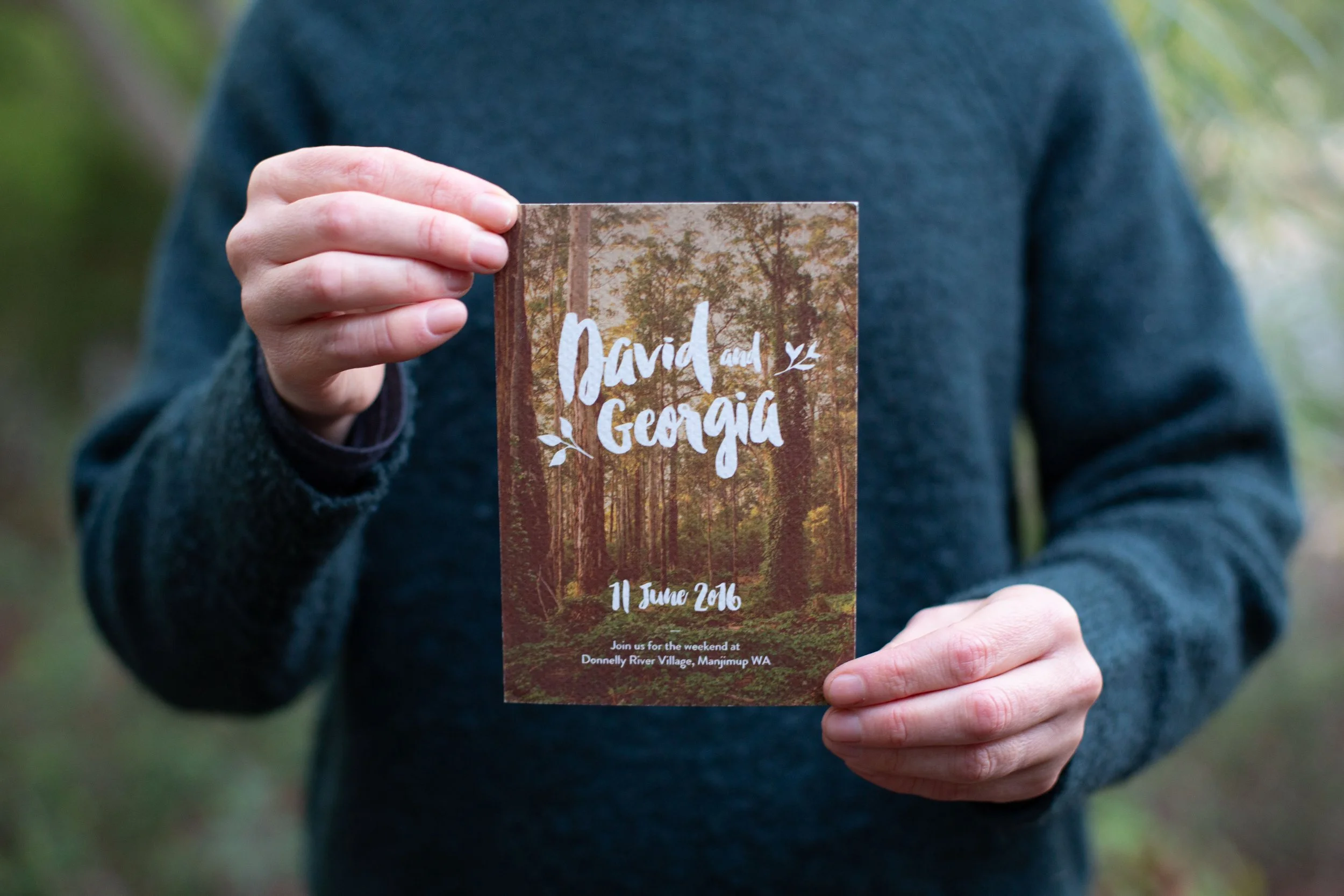 Person holding a wedding invitation with a forest background that reads "David and Georgia, 11 June 2016," with additional details about the location at Donnelly River Village, Manjimup WA.