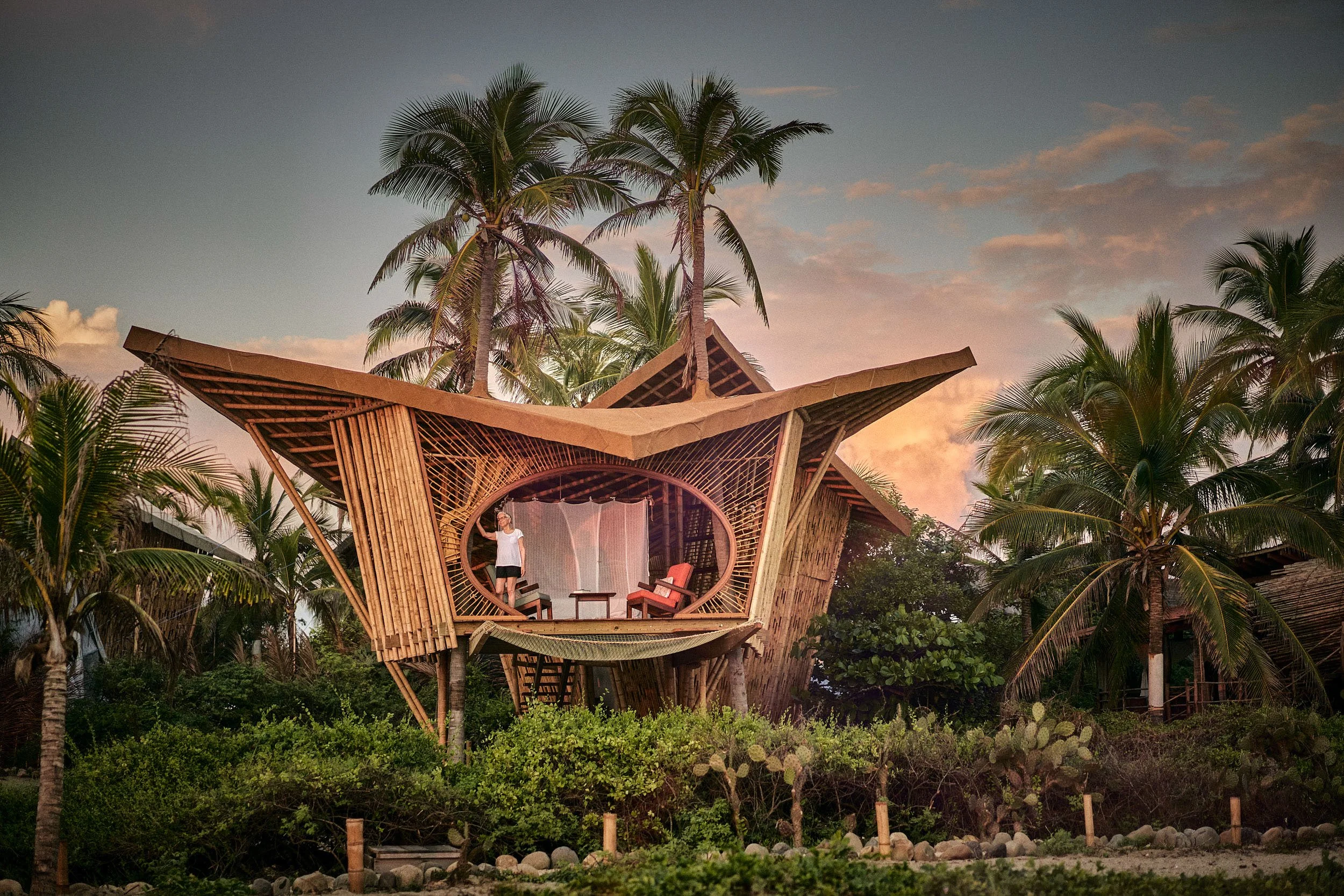 A unique wooden house on stilts surrounded by lush greenery and tall palm trees, with a sunset sky in the background.