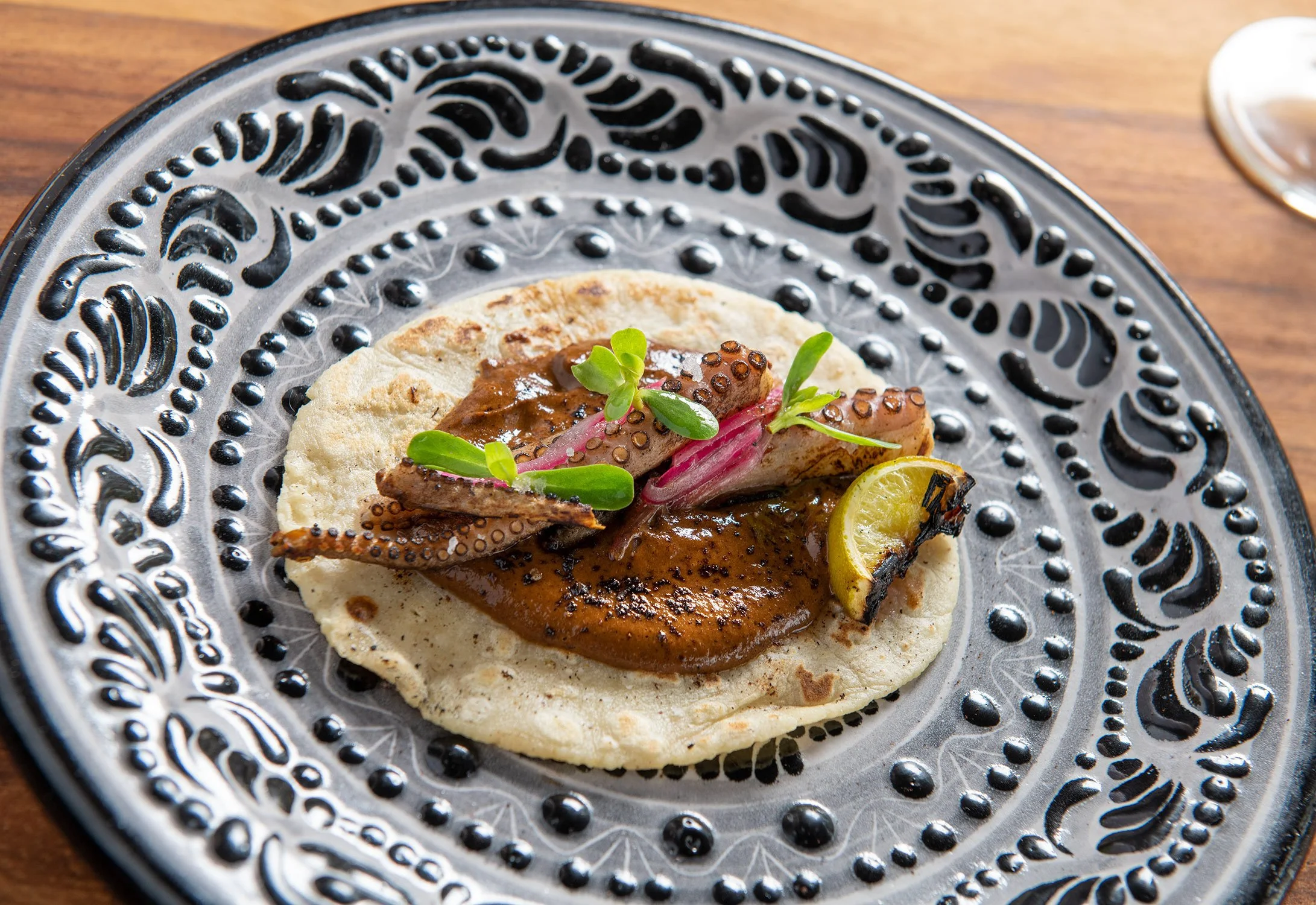 A traditional Mexican taco on a decorative black and silver plate, topped with grilled octopus, garnished with pink onions, a lemon wedge, and fresh green herbs, with a glass of white wine in the background.