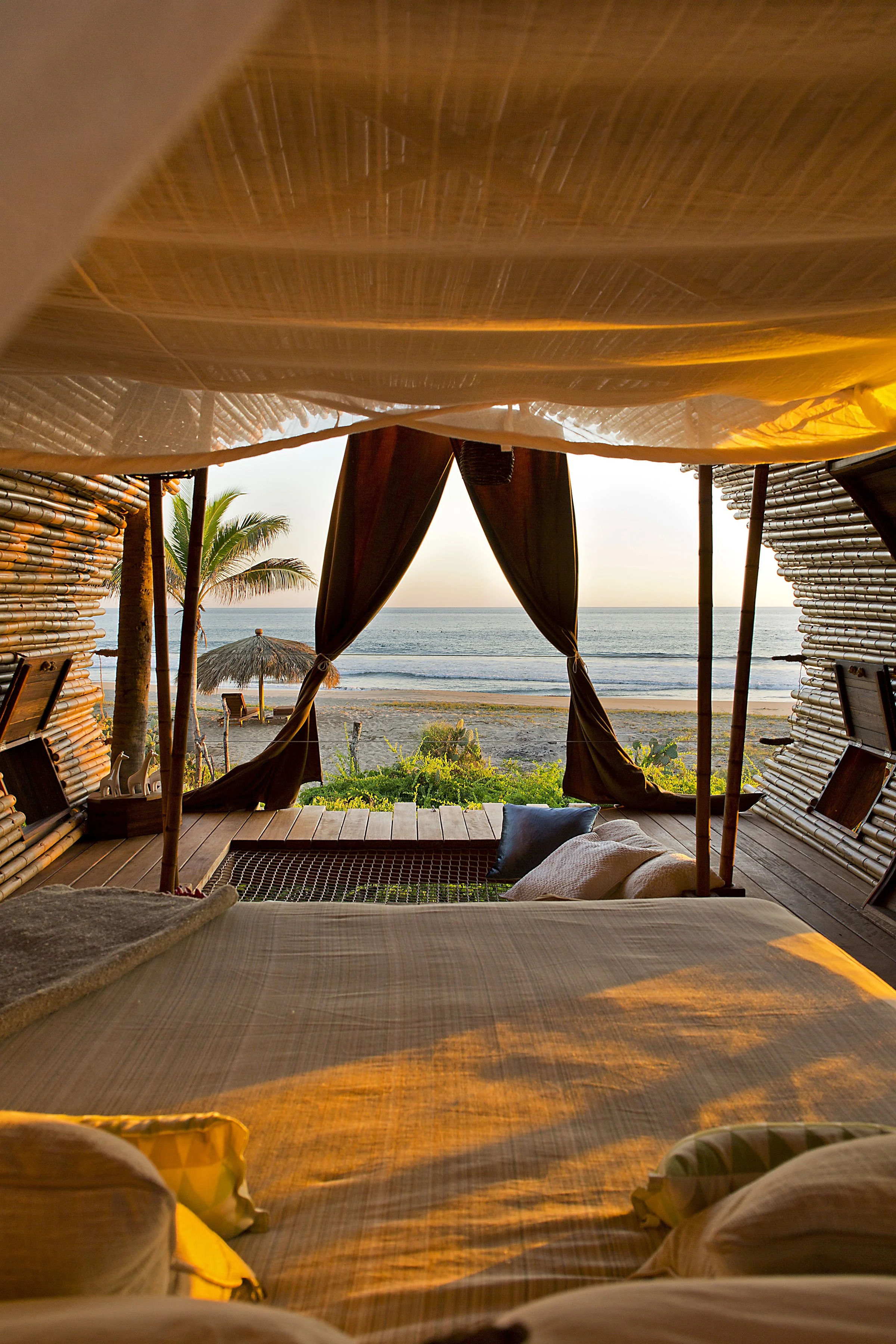 View from inside a bungalow overlooking the beach at sunset, with a bed and pillows in the foreground, curtains open to a sandy beach, palm trees, and the ocean beyond.