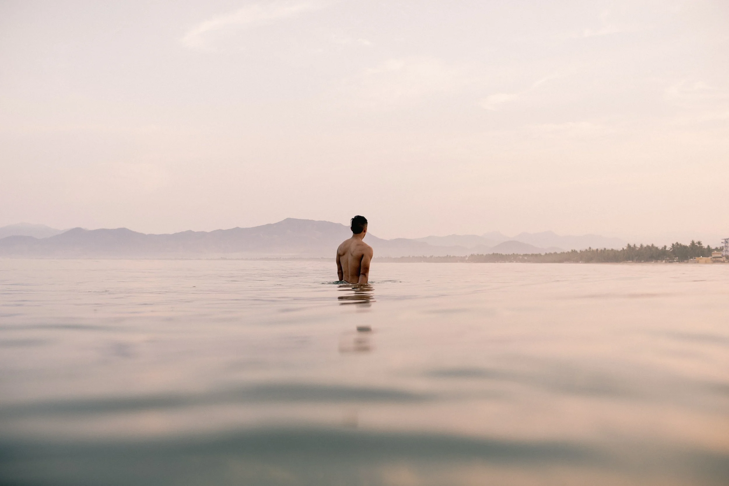 A person standing in calm water with distant mountains and a hazy sky in the background.