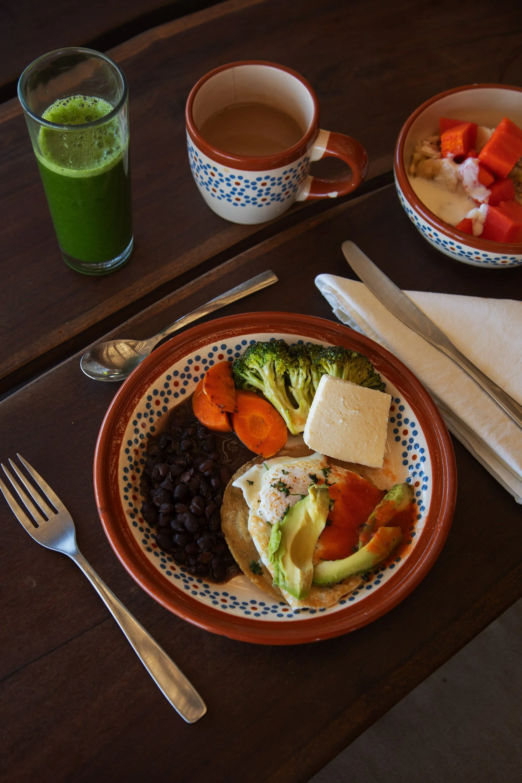 A breakfast plate with black beans, broccoli, carrots, a slice of cheese, and avocado on a dark wooden table. A spoon and knife are on napkins to the right. In the background, a glass of green juice, a cup of coffee, and a bowl of fruit with watermel