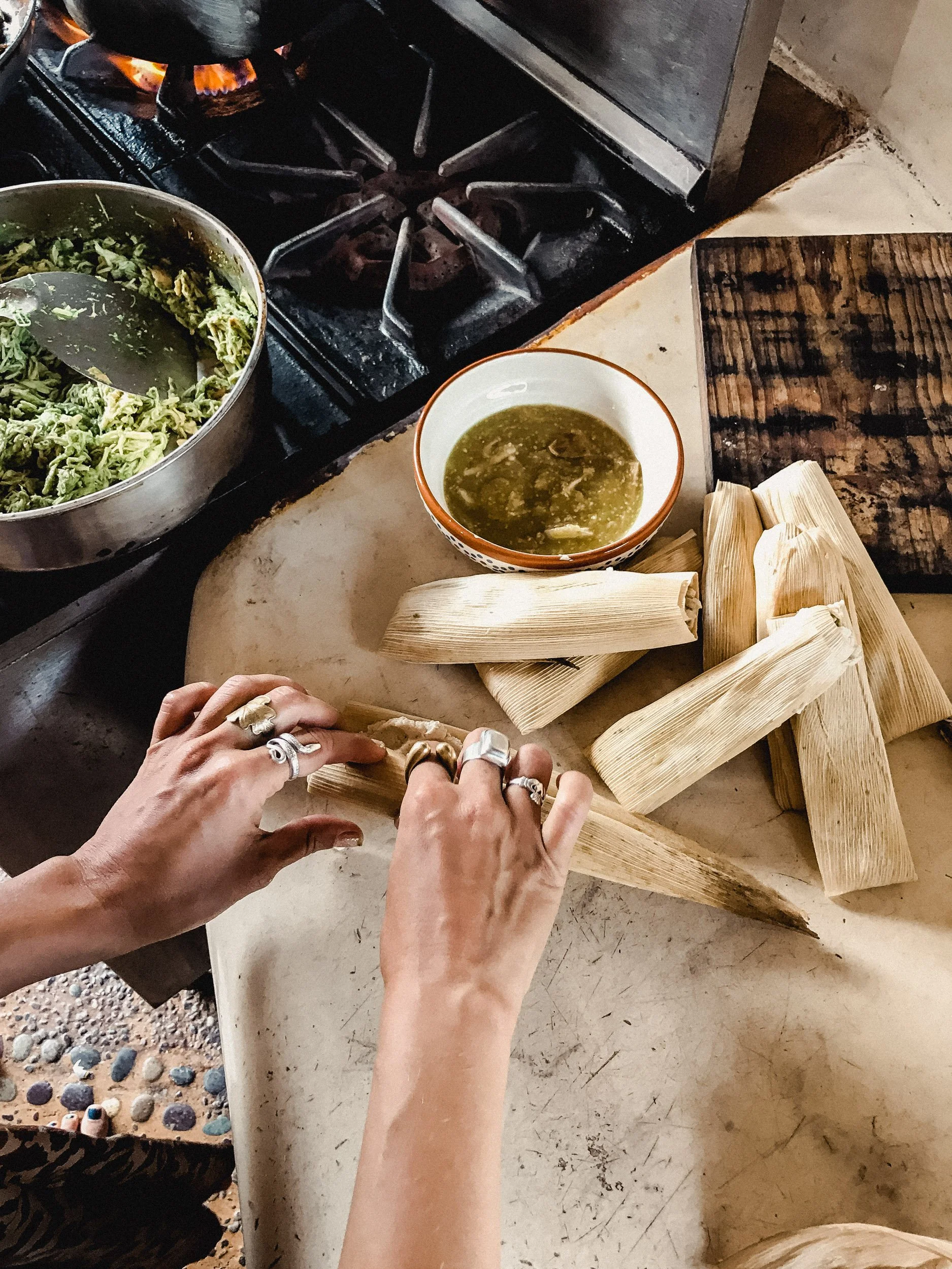 Hands preparing tamales on a kitchen counter, with a bowl of green sauce, a bunch of raw corn husks, and a stove with a pot of greens nearby.