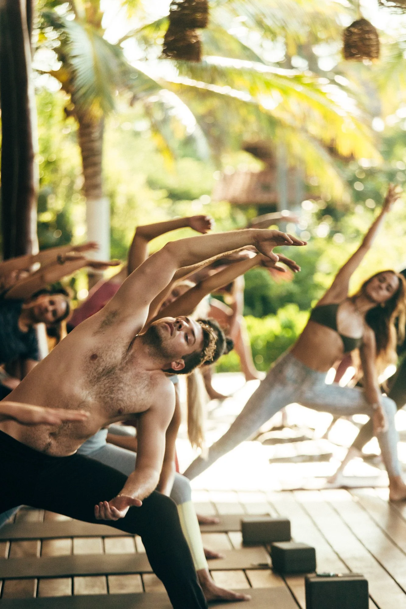 Group of people doing yoga outdoors in a tropical setting, stretching and practicing poses on mats.