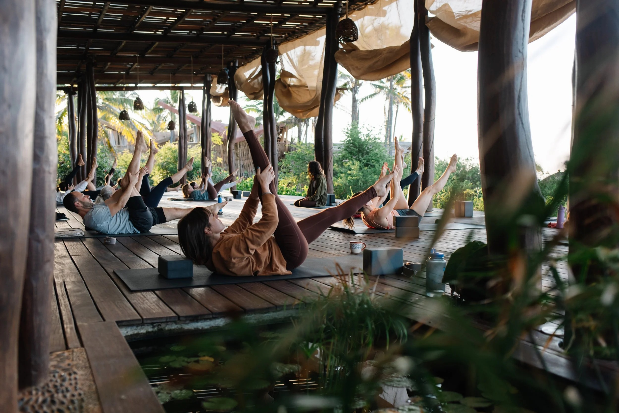 People practicing yoga on wooden deck with large windows and greenery outside, during sunrise or sunset.