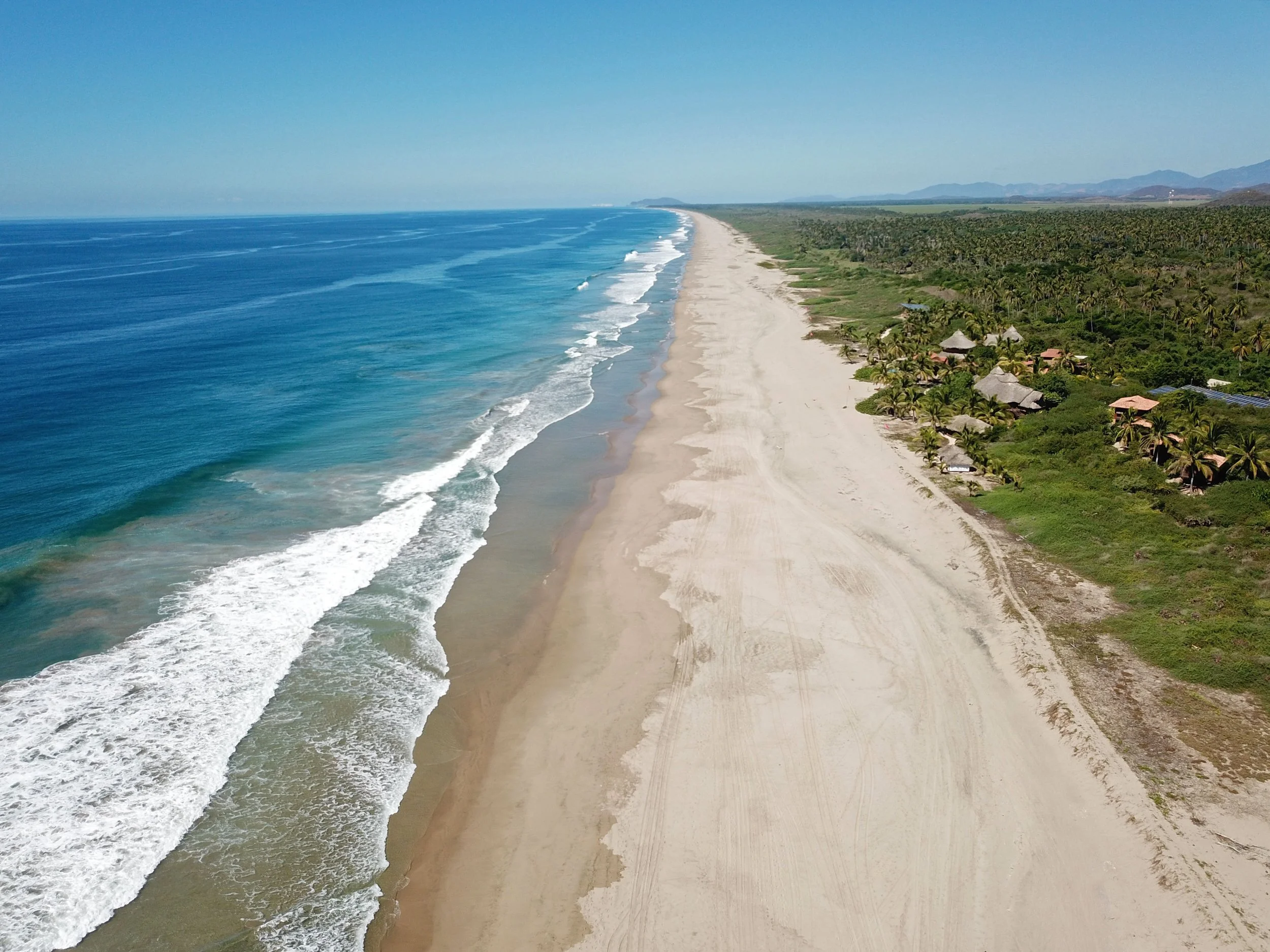 Aerial view of a long, sandy beach with turquoise ocean waves on the left and green vegetation with scattered houses on the right, extending into the horizon.
