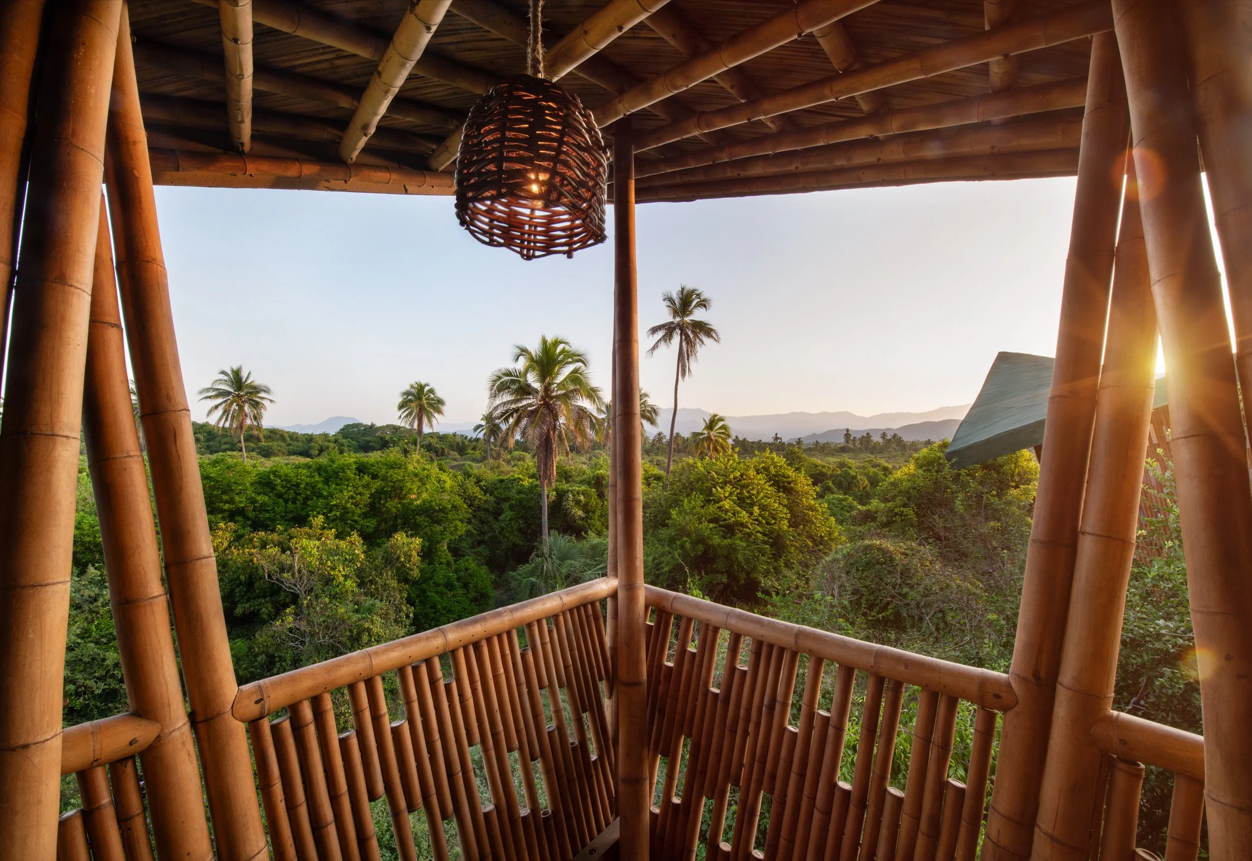 Bamboo balcony overlooking a lush tropical forest with palm trees, mountains in the distance, and sunlight.