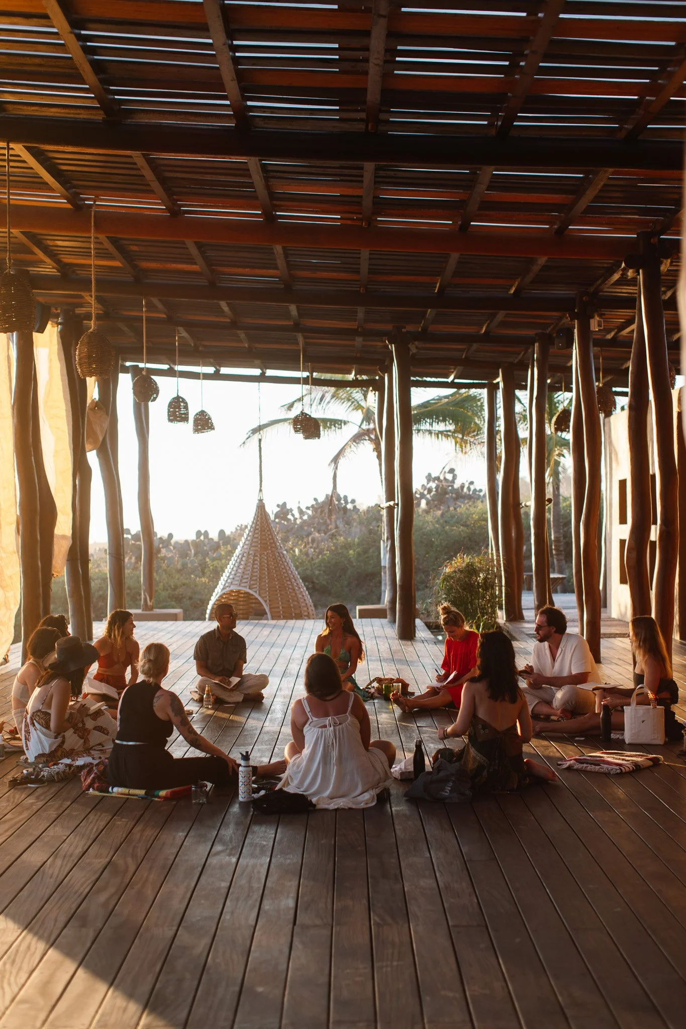 Group of people sitting cross-legged in a circle on wooden deck under a rustic, open-air roof, with palm trees and sunset in the background.