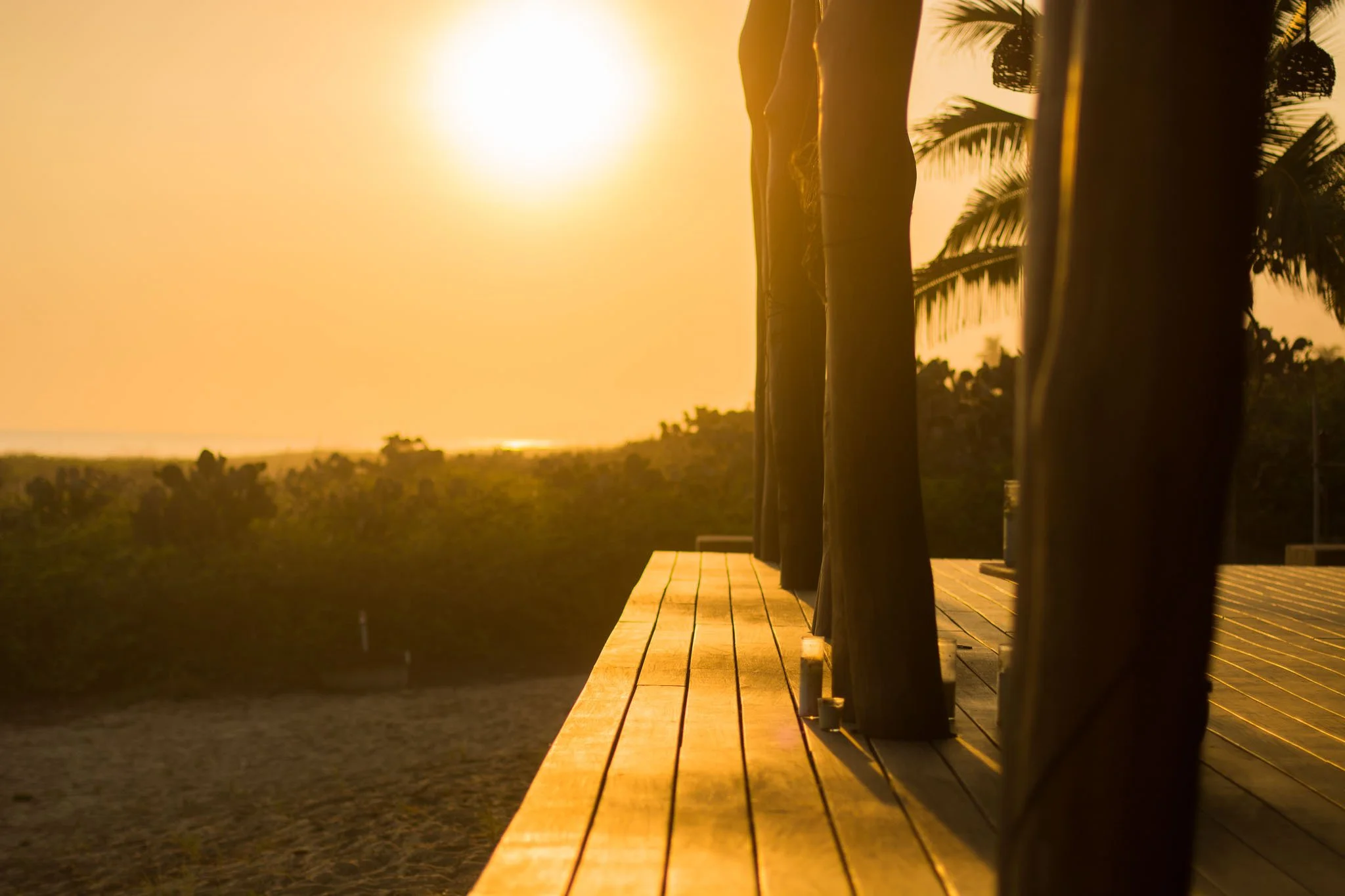 View of a wooden deck with railing, overlooking a beach and distant trees, during sunset or sunrise with the sun low in the sky.