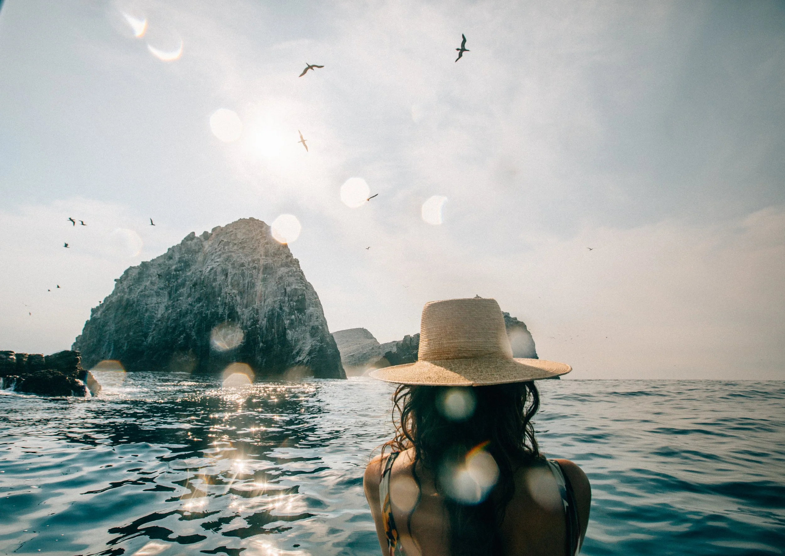A woman wearing a wide-brimmed straw hat is sitting in the water facing the ocean, with cliffs and a large rock formation in the distance, and birds flying in the sky.